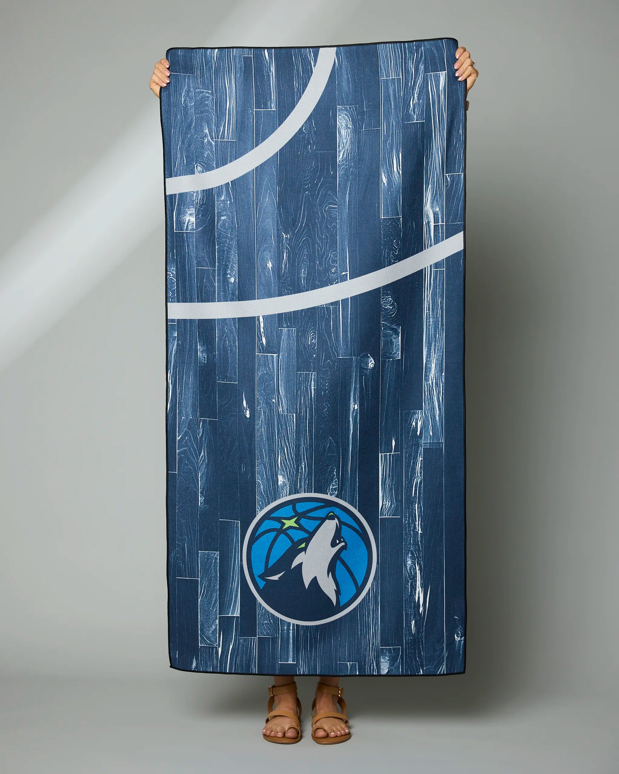 A person holds up the Geometry Timberwolves Zone towel, which is rectangular with a wood-like blue pattern, white court lines, and a basketball logo featuring a wolf howling at a green star near the center bottom.