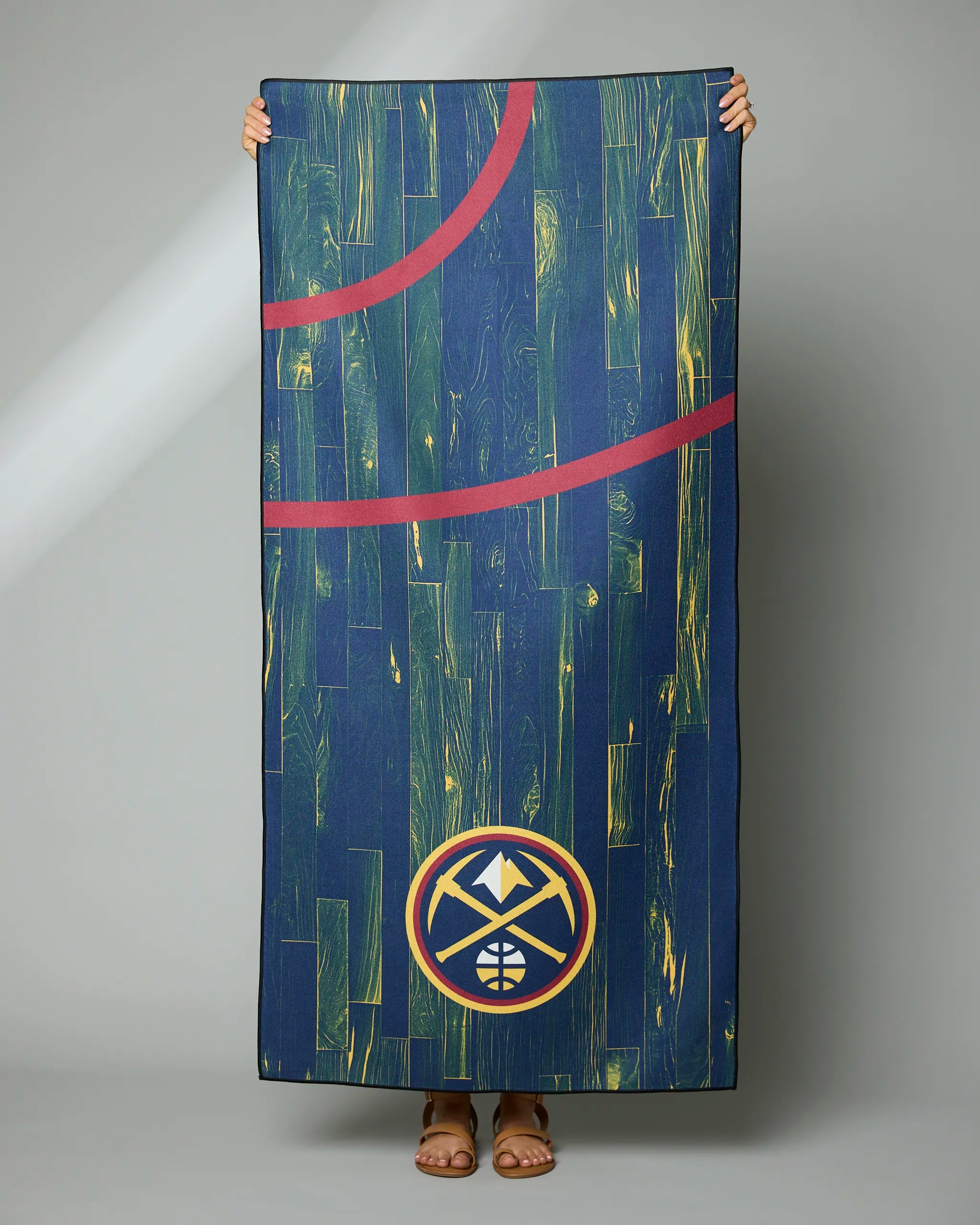 A person holds the Geometry Nuggets Zone towel featuring a green and yellow basketball court design with red lines and the Denver Nuggets logo; only their hands and feet are visible.