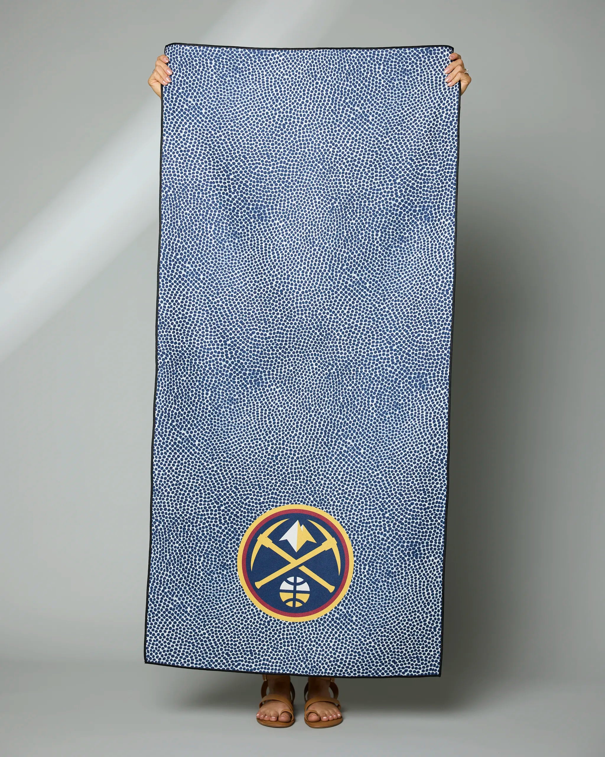 Someone holds up the Geometry Nuggets Grip, a large blue towel with a dotted pattern and the Denver Nuggets logo near the bottom center; only their hands and feet are visible.