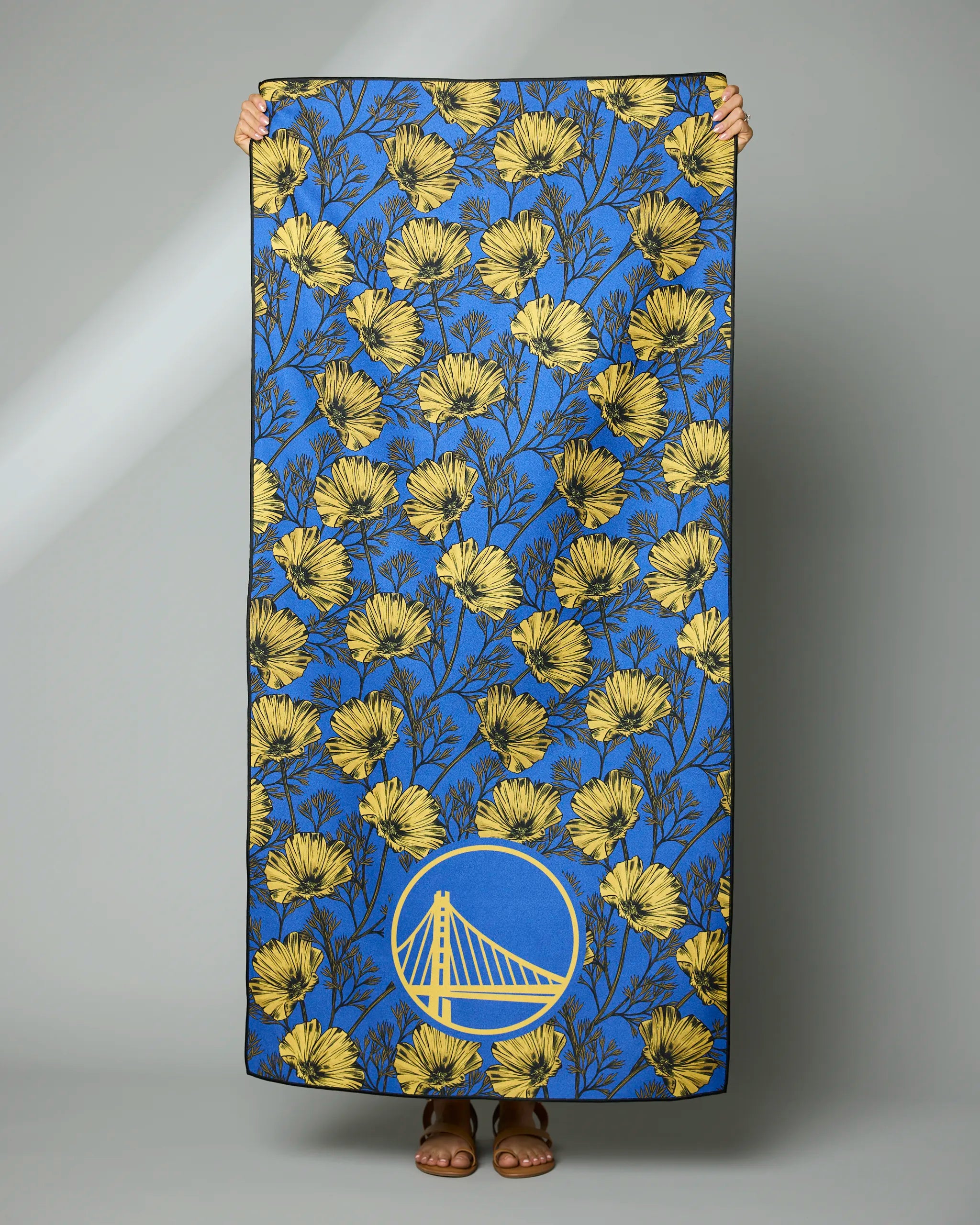 A person holds up the Geometry Warriors Floral towel, which has a blue background with yellow flowers and a yellow Golden State Warriors bridge logo at the bottom center. Only their hands and feet are visible.