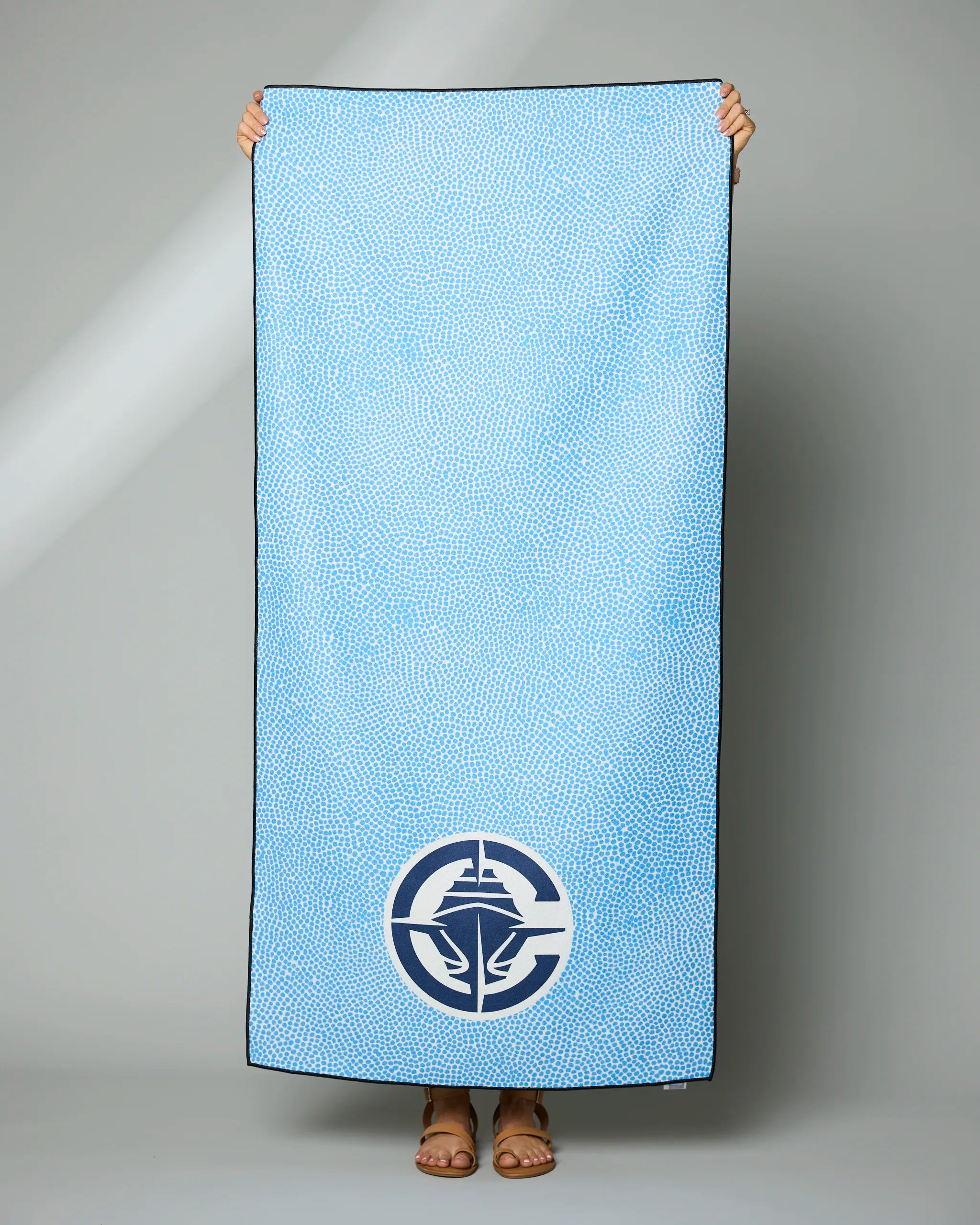 A person holds up the Geometry Clippers Grip towel, a rectangular light blue towel with small white dots, a dark navy border, and a large circular white and navy emblem with a stylized leaf near the bottom.