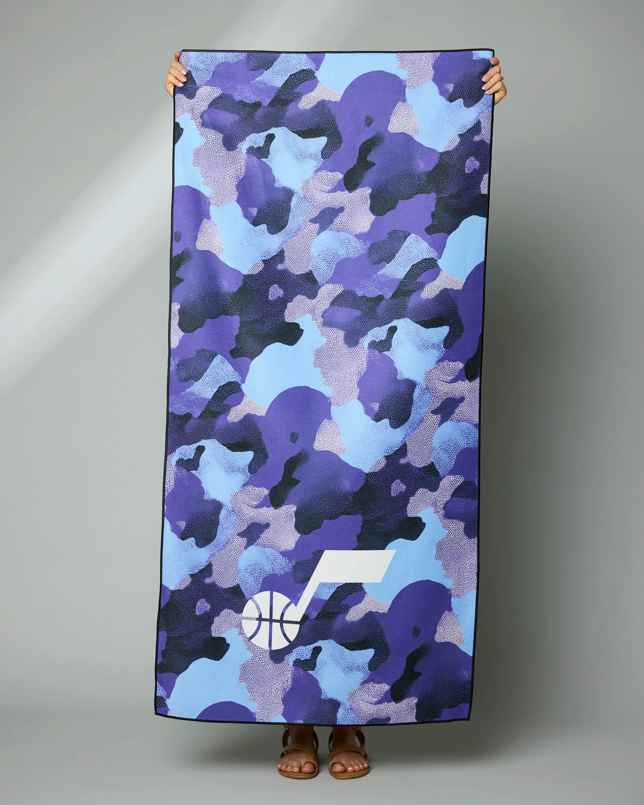 A person holds up the Geometry Jazz Camo towel, featuring a blue, purple, and black camouflage design with a white basketball and music note logo at the bottom. Only their hands and feet are shown.