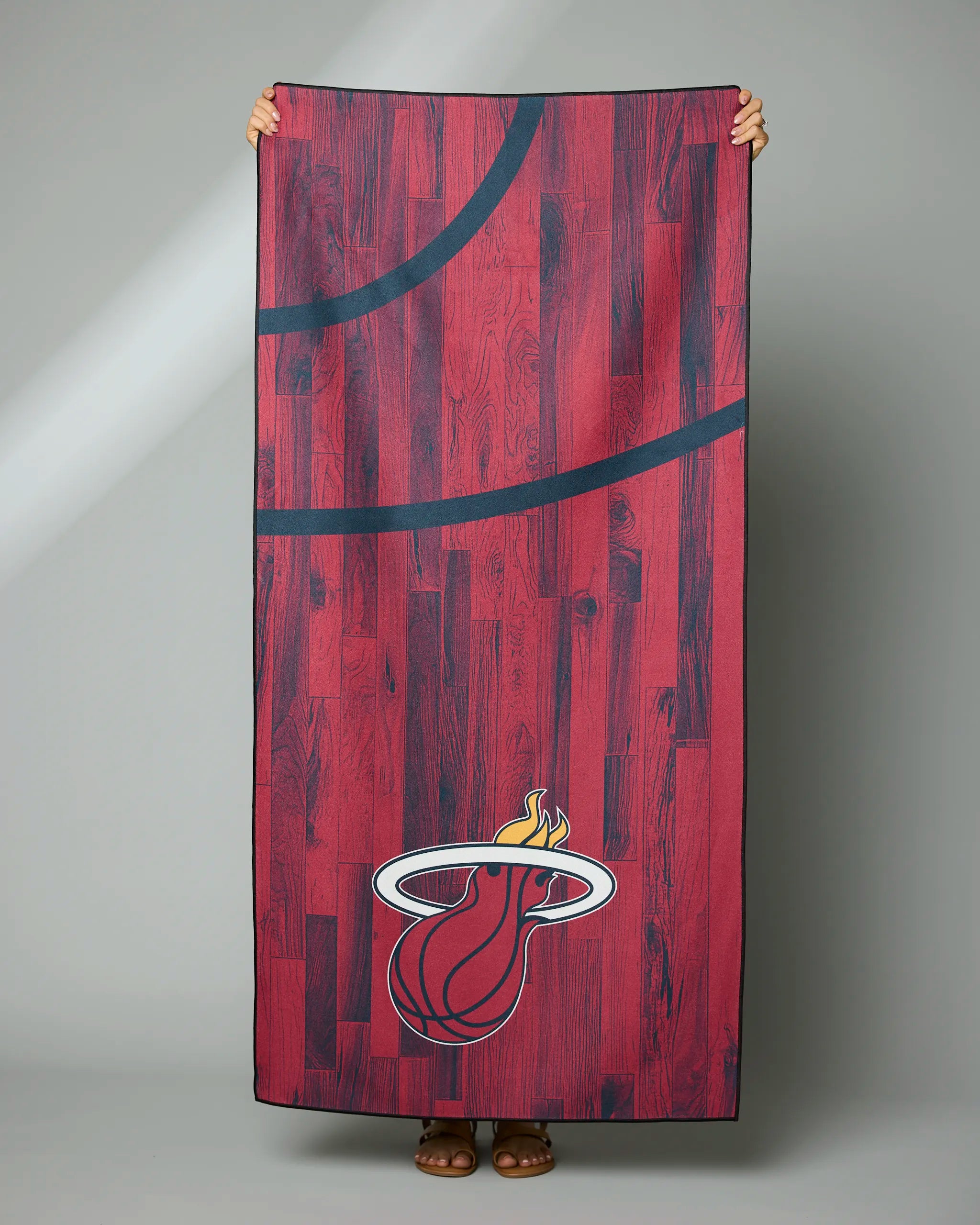 Someone holds up Geometrys Heat Zone towel, which is red with a wood floor pattern and displays the stylized Miami Heat logo with flames and black lines like a basketball court.