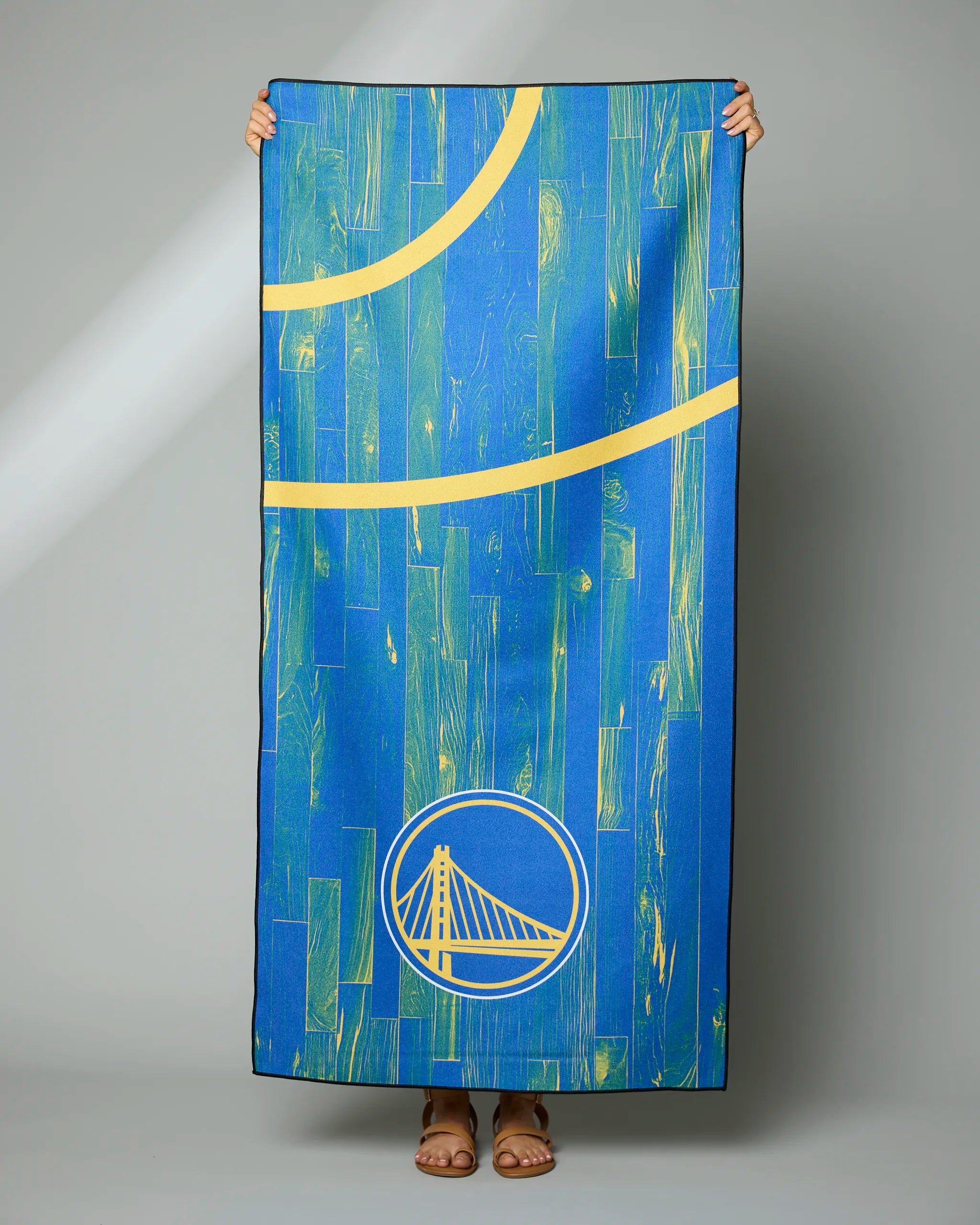 A person is holding up the Warriors Zone towel by Geometry, featuring a blue and yellow basketball court design with the Golden State Warriors logo. Only their hands and feet are visible against a plain gray background.