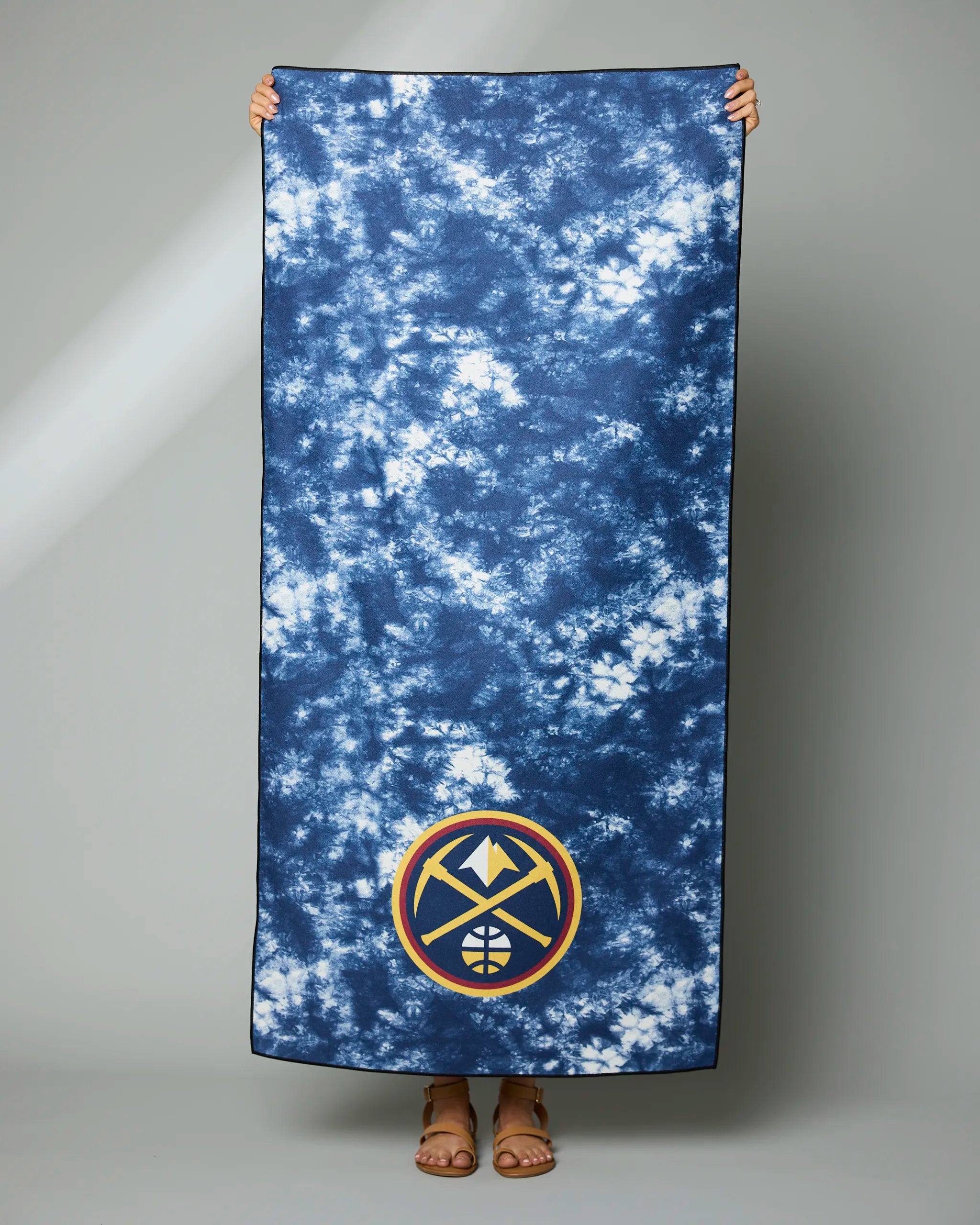 A person holds up the large Geometry Nuggets Flash towel featuring a blue and white tie-dye design and a Denver Nuggets logo in the corner, with only their hands, feet, and sandals showing behind it.