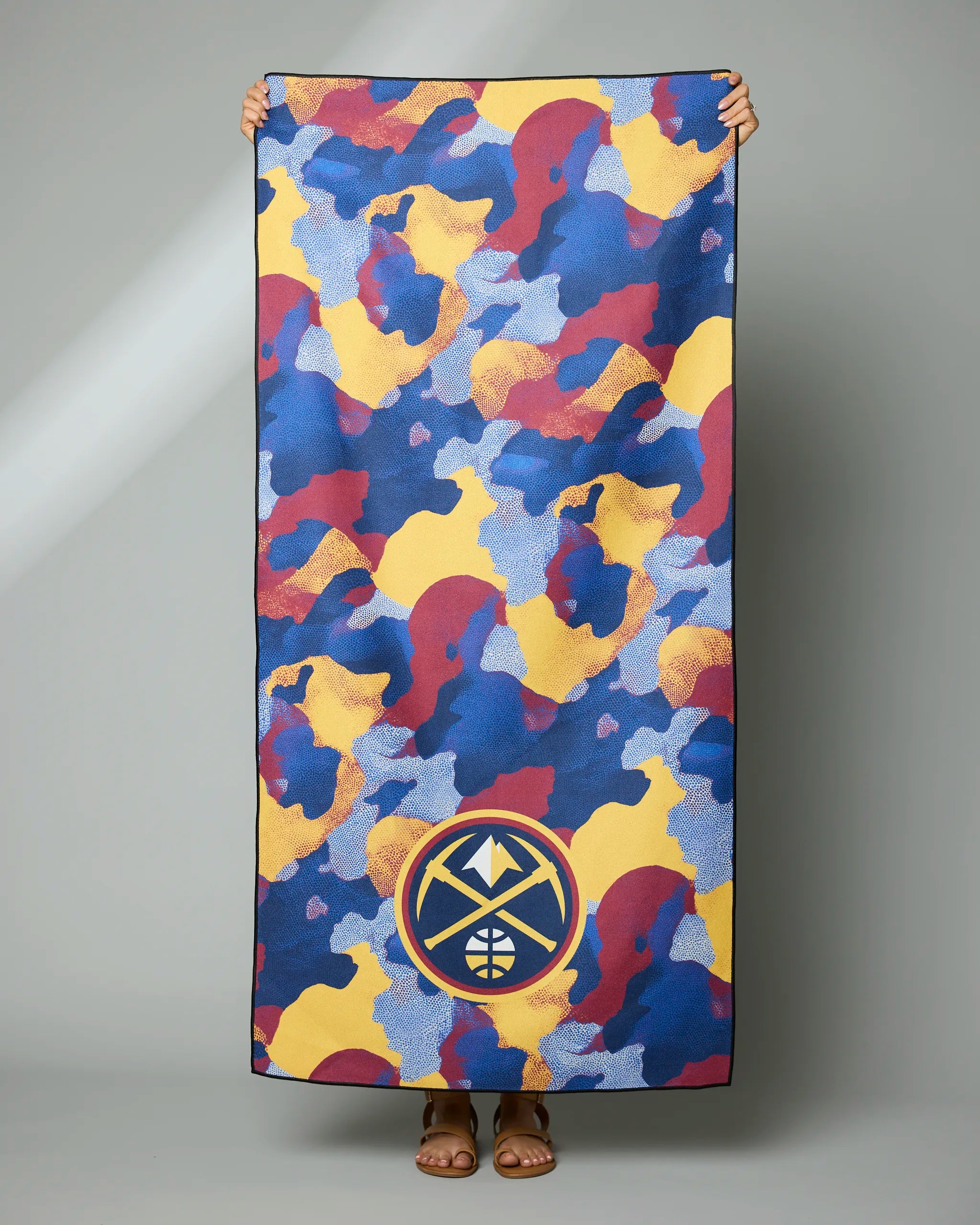 A person holds the Geometry Nuggets Camo towel, featuring a rectangular shape, abstract blue, yellow, and maroon camouflage design, and a Denver Nuggets logo near the bottom. Only their hands, feet, and sandals are visible.