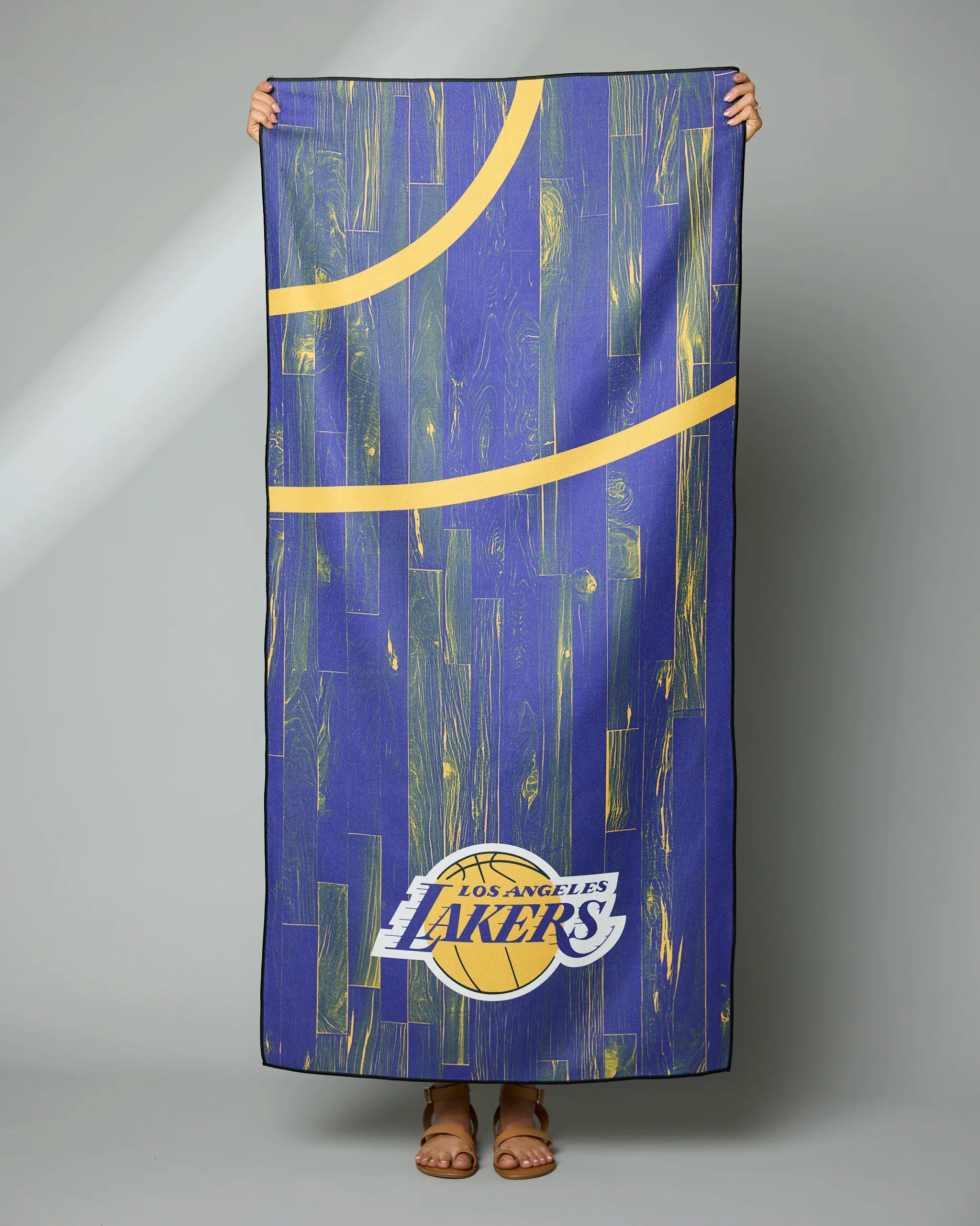 A person holds the Geometry Lakers Zone rectangular towel, which has a blue and yellow wood grain pattern, partial yellow court lines, and the Los Angeles Lakers logo. The persons face is not shown.