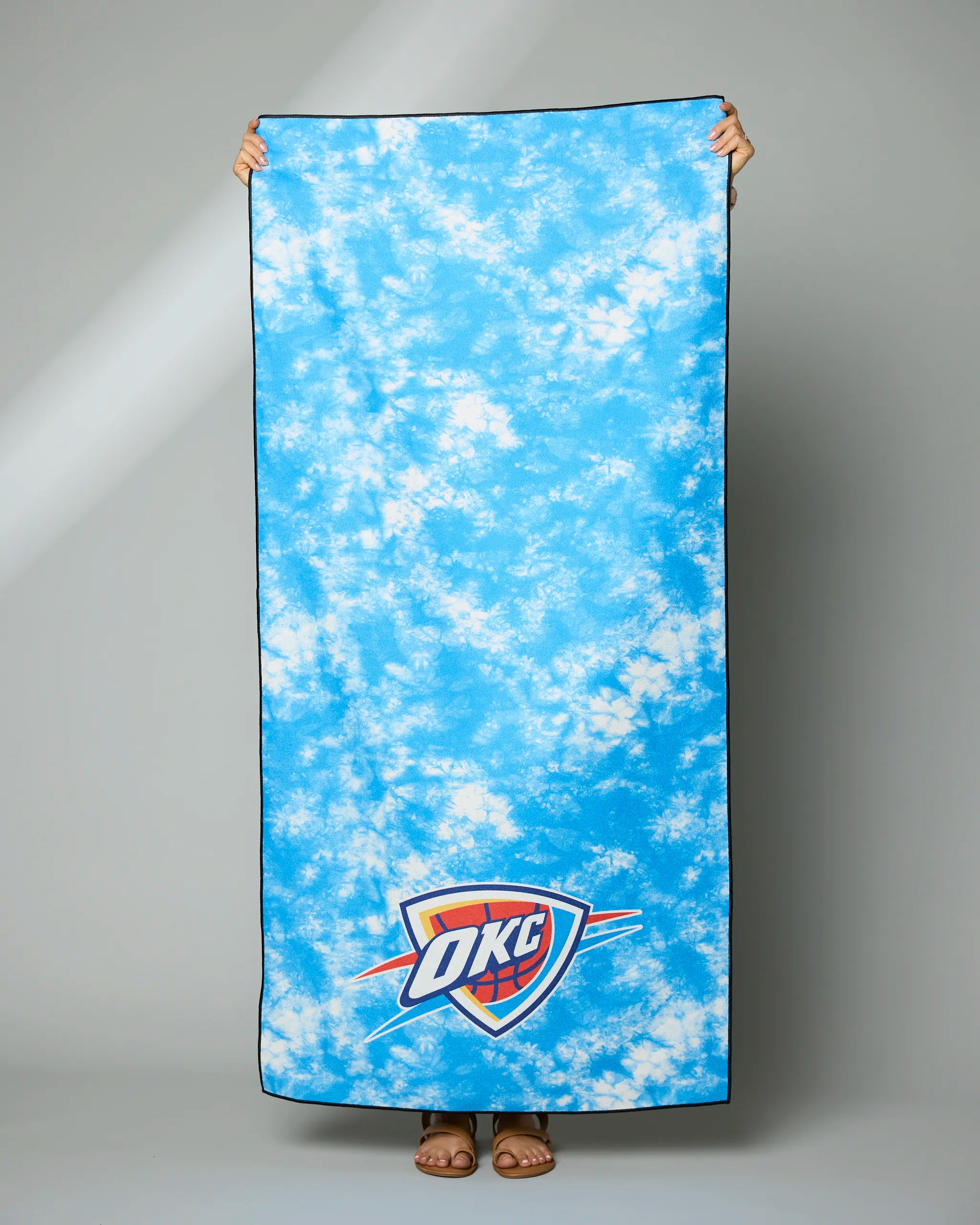 A person holds up the Geometry OKC Flash towel, a large blue rectangular towel with white cloud patterns and the Oklahoma City Thunder logo near the bottom. Only their hands and feet are visible.