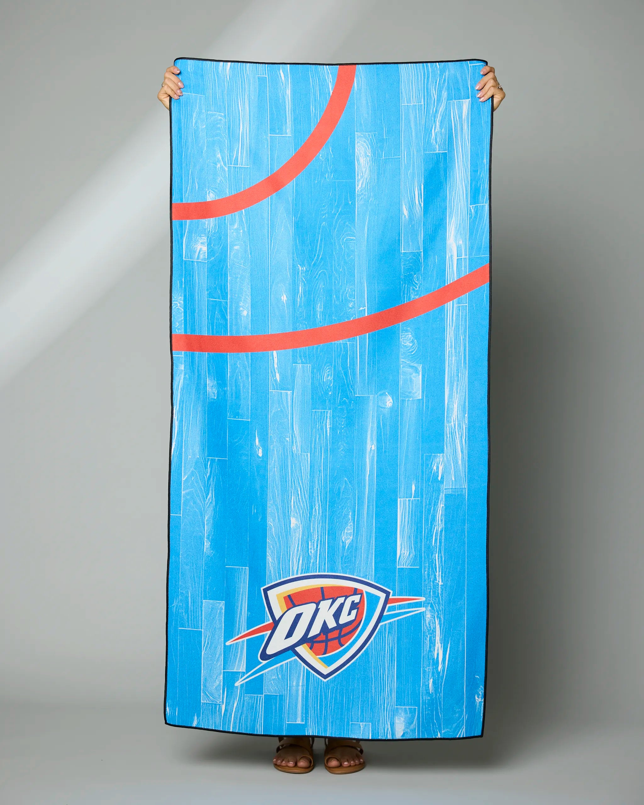 A person holds up the Geometry OKC Zone towel, which is rectangular, blue, and features a hardwood court design with red basketball lines and the Oklahoma City Thunder logo at the bottom—only their hands and feet are visible.