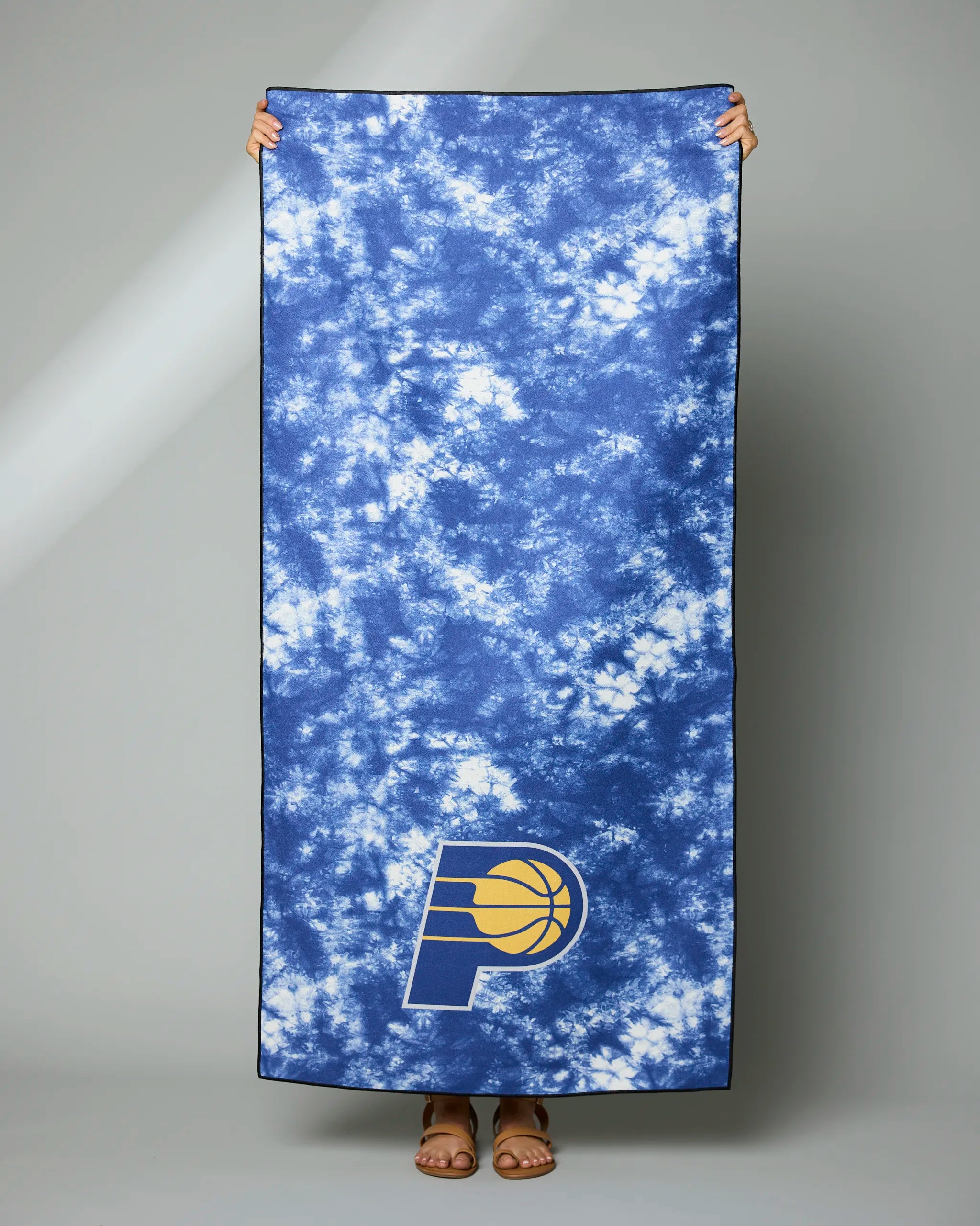 A person holding up the Geometry Pacers Flash towel, a blue and white tie-dye design with the Indiana Pacers logo at the bottom. Only their hands and feet are visible.