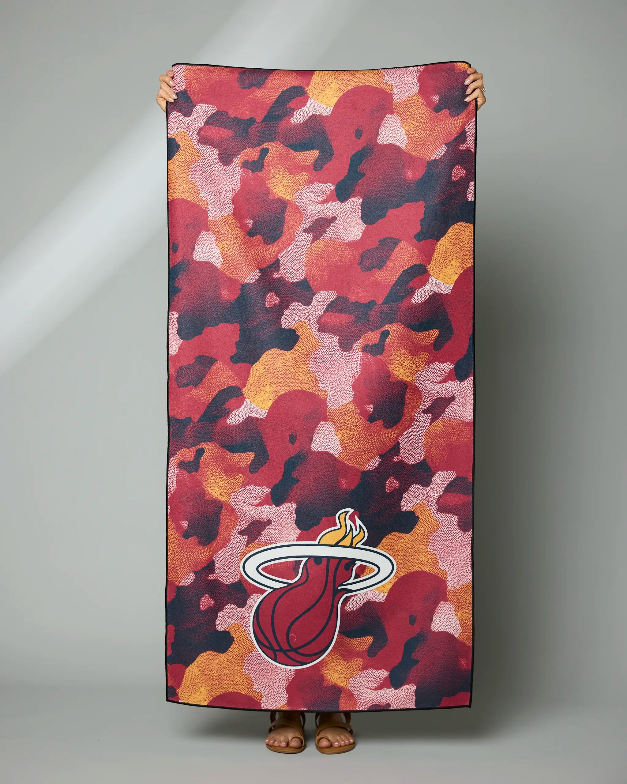 A person holds the Geometry Heat Camo towel, featuring a red, orange, and pink camouflage design and the Miami Heat logo at the bottom. Only their hands and feet are visible.