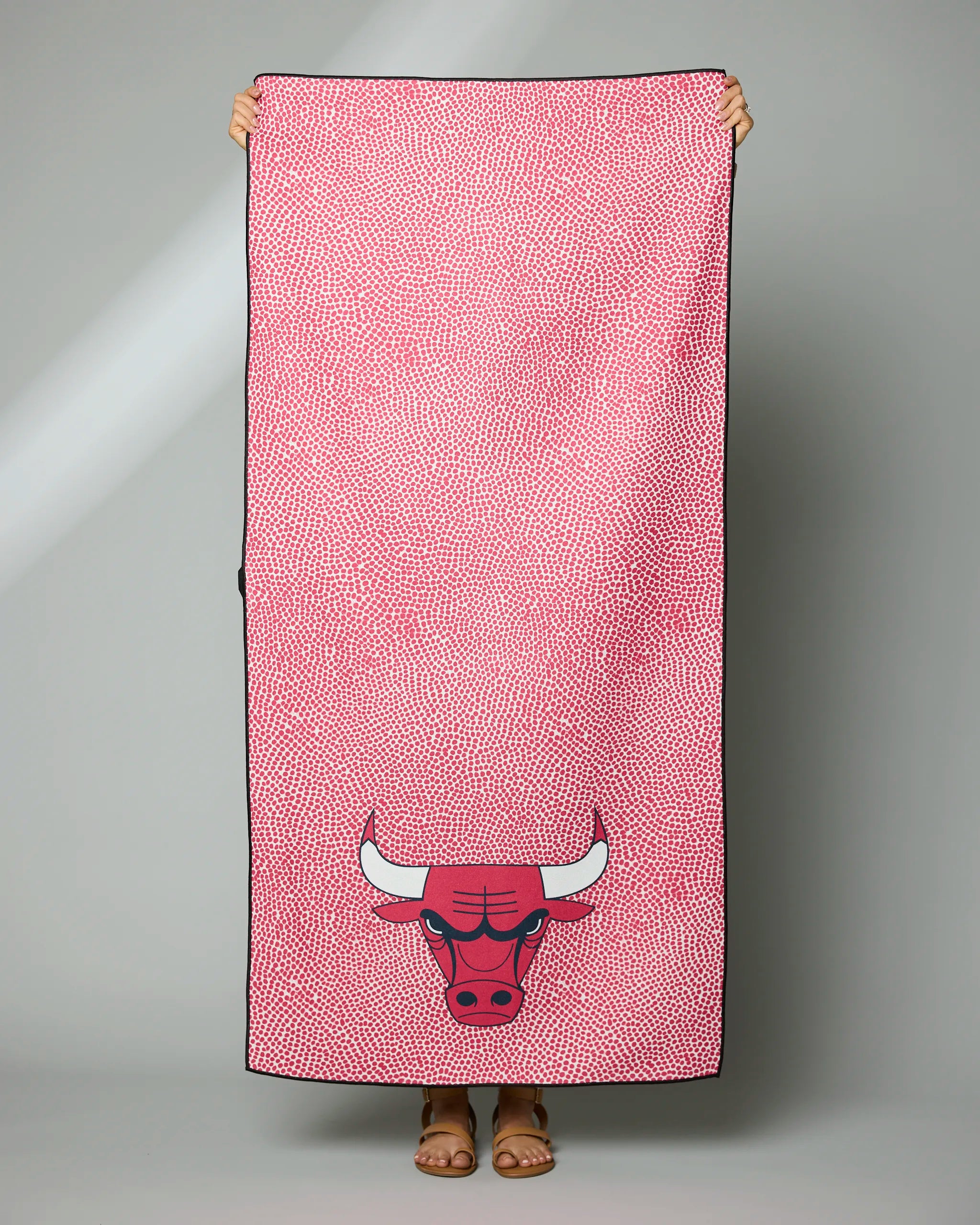 A person stands holding the Geometry Bulls Grip towel, a pink towel with small white dots and a large red Chicago Bulls bull head logo at the bottom, hiding their face behind it.