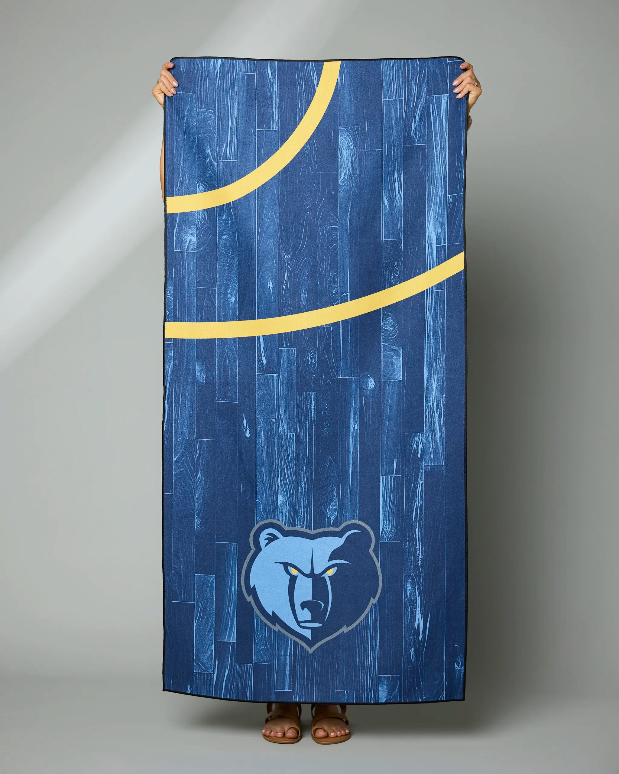 Against a plain gray background, a person displays the Geometry “Grizzlies Zone” towel—a rectangular blue towel with yellow basketball court lines and the Memphis Grizzlies bear logo at the bottom.