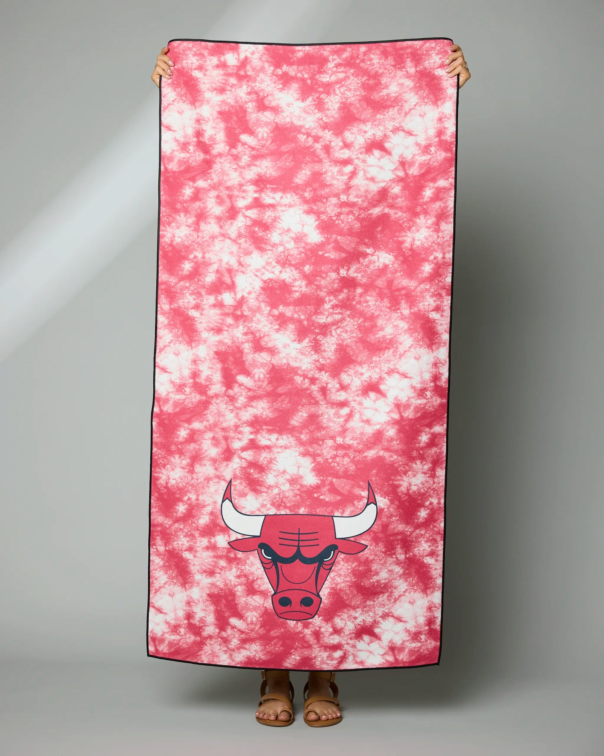 A person holds up the Geometry Bulls Flash towel, featuring a pink and white tie-dye design with the Chicago Bulls logo at the bottom. Only their hands and feet can be seen behind it.