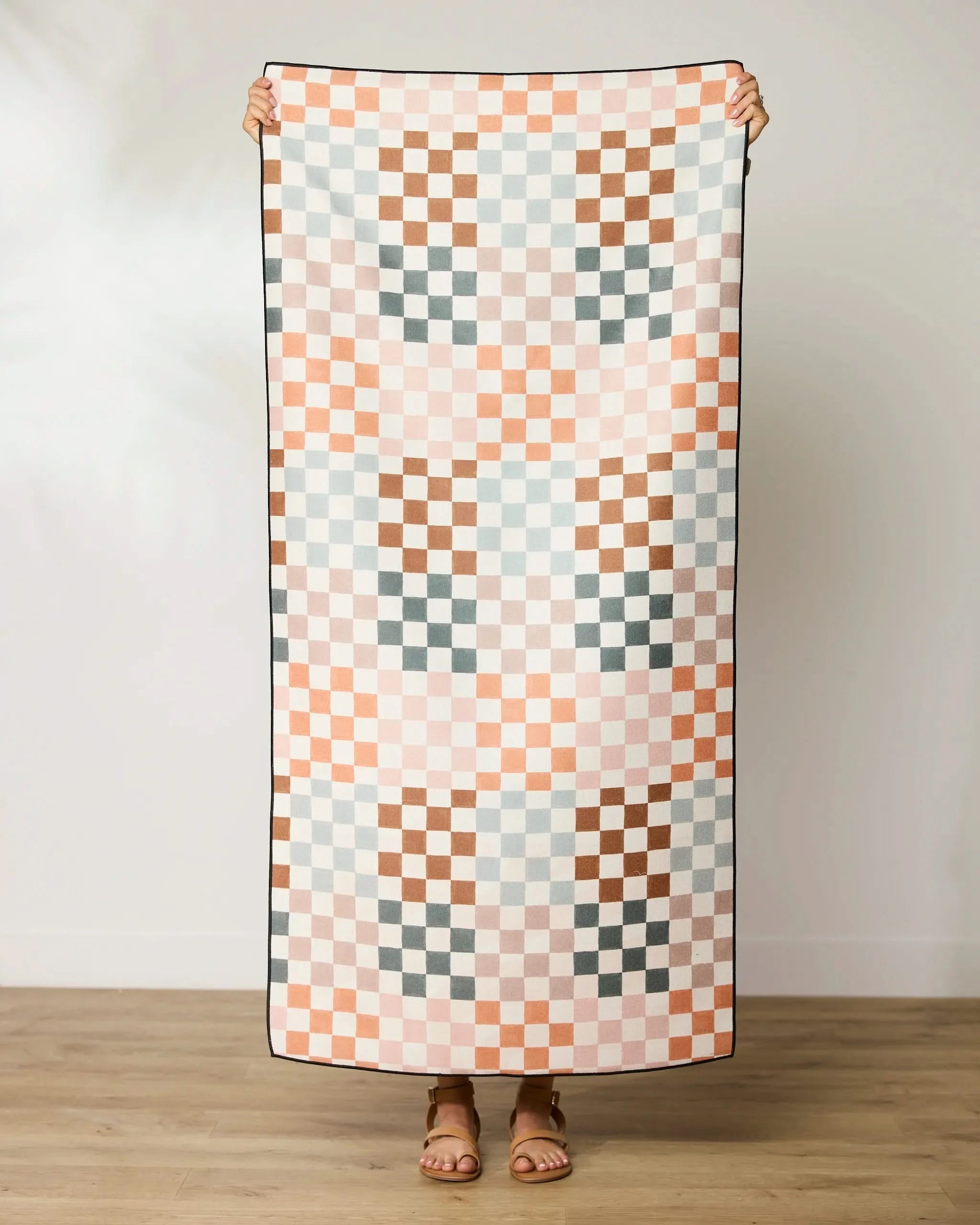 A person stands holding up the GEOMETRY Autumn Checkers micro-loop beach towel, featuring a pastel pink, peach, brown, and light blue checkerboard pattern. Their head is not visible and they wear sandals on a wooden floor.