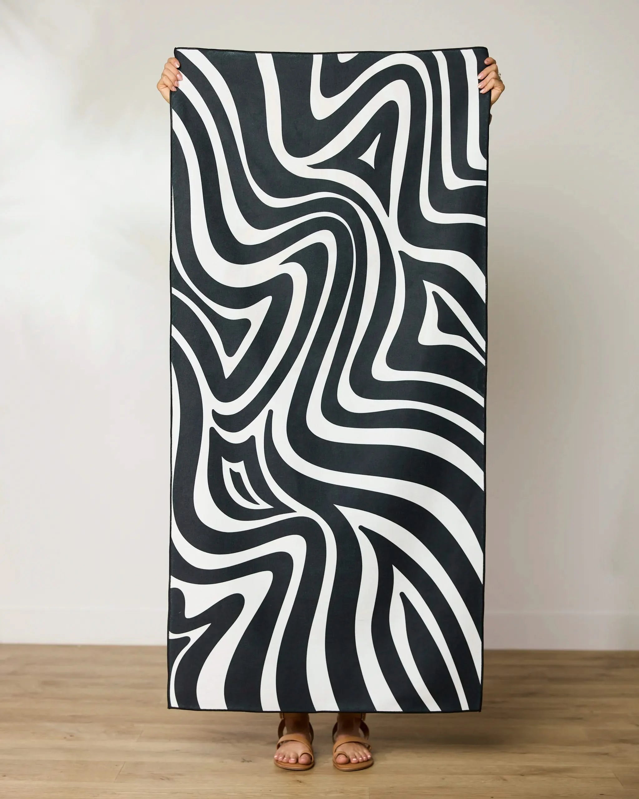 A person stands on a wooden floor, holding up the GEOMETRY Dizzy Black quick-drying beach towel featuring bold black and white wavy abstract patterns that create an optical illusion. Their face and upper body are hidden behind the towel.