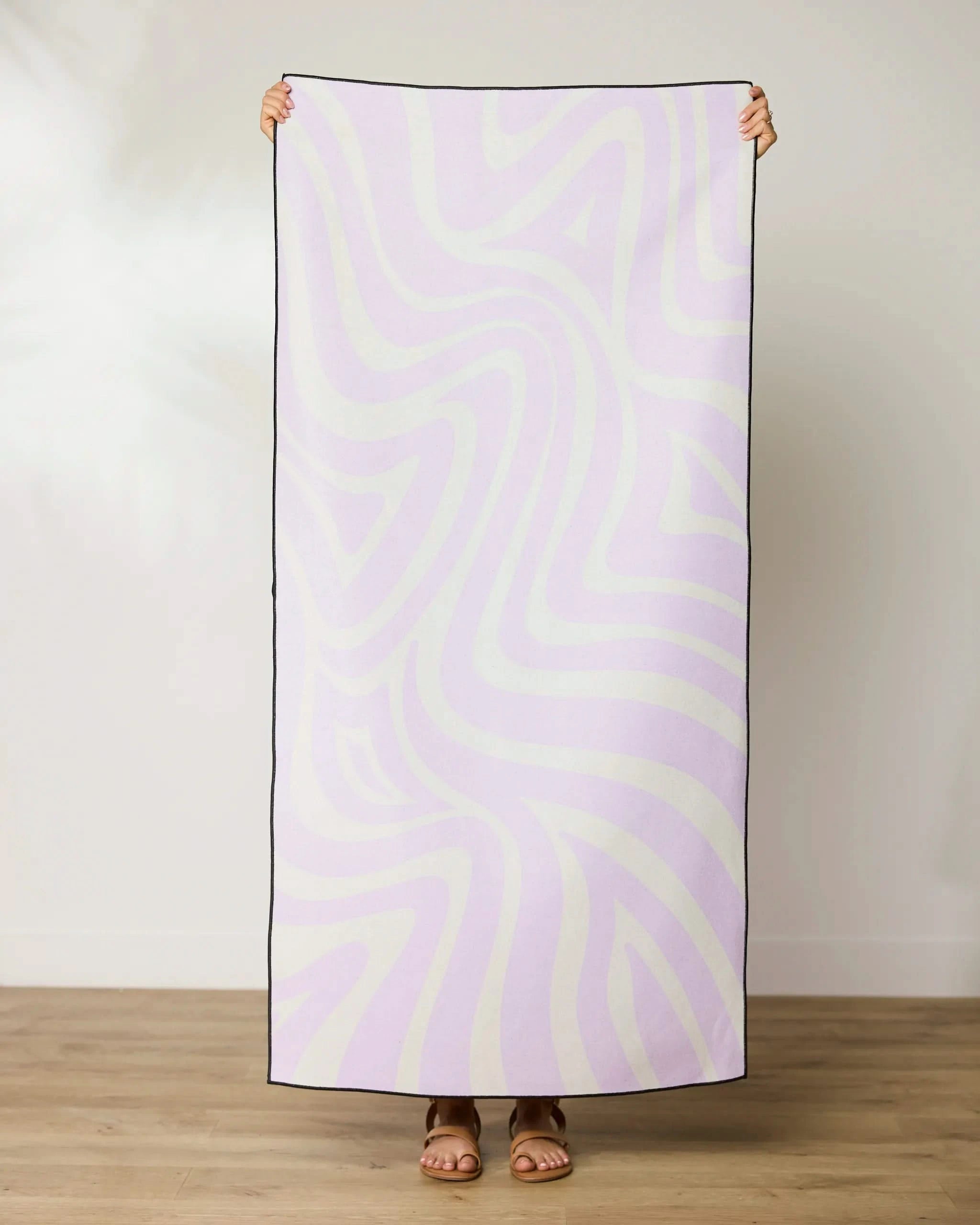 A person stands on a wooden floor, holding up the GEOMETRY Dizzy Lilac towel—made from recycled materials with a pastel purple and white wavy design. Only their hands and sandaled feet are seen behind the towel.