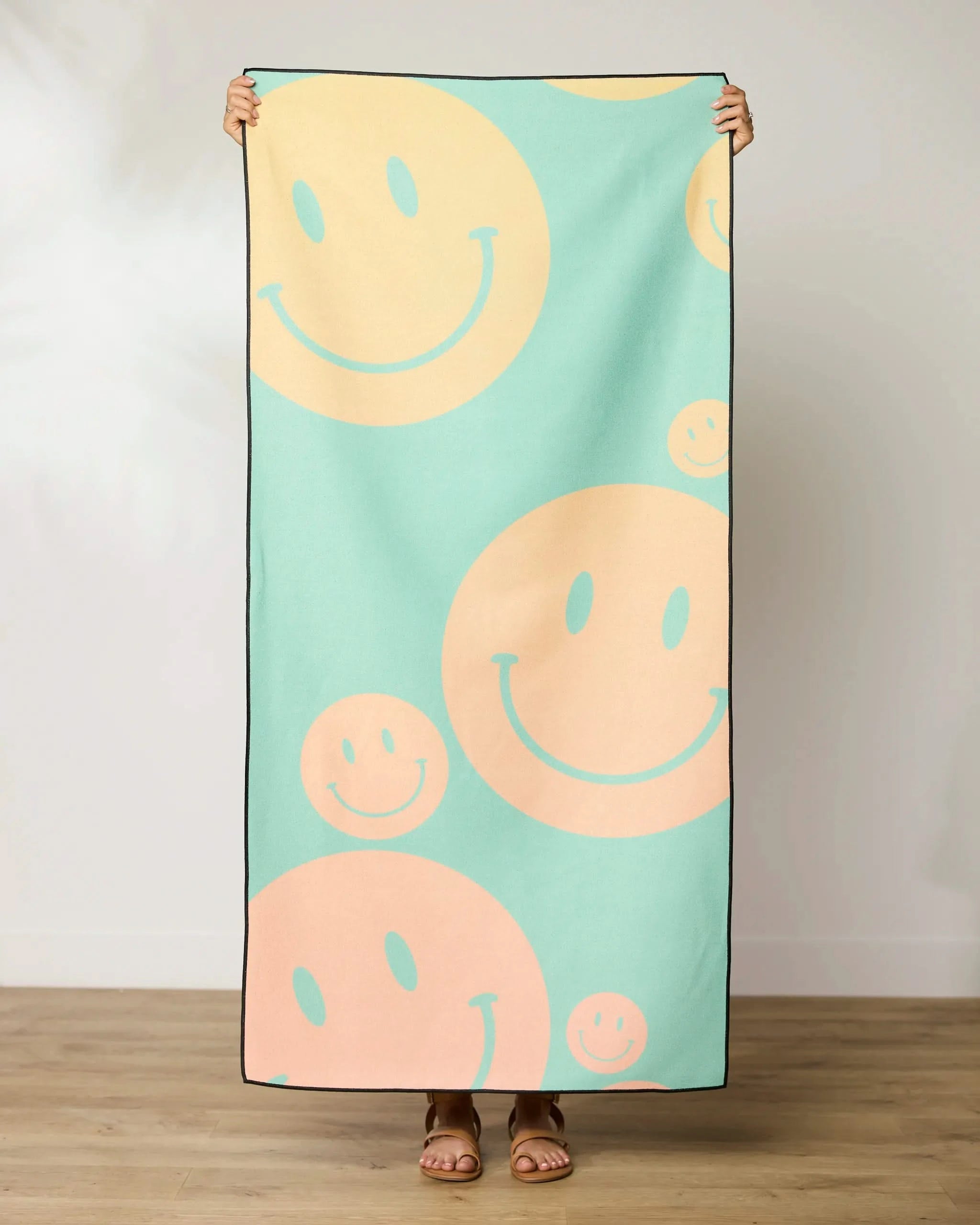 A person stands indoors holding up the GEOMETRY Happy Days Micro-loop Beach Towel, featuring a pastel green background with large peach and yellow smiley faces. Only their hands and feet are visible. Made from recycled materials.