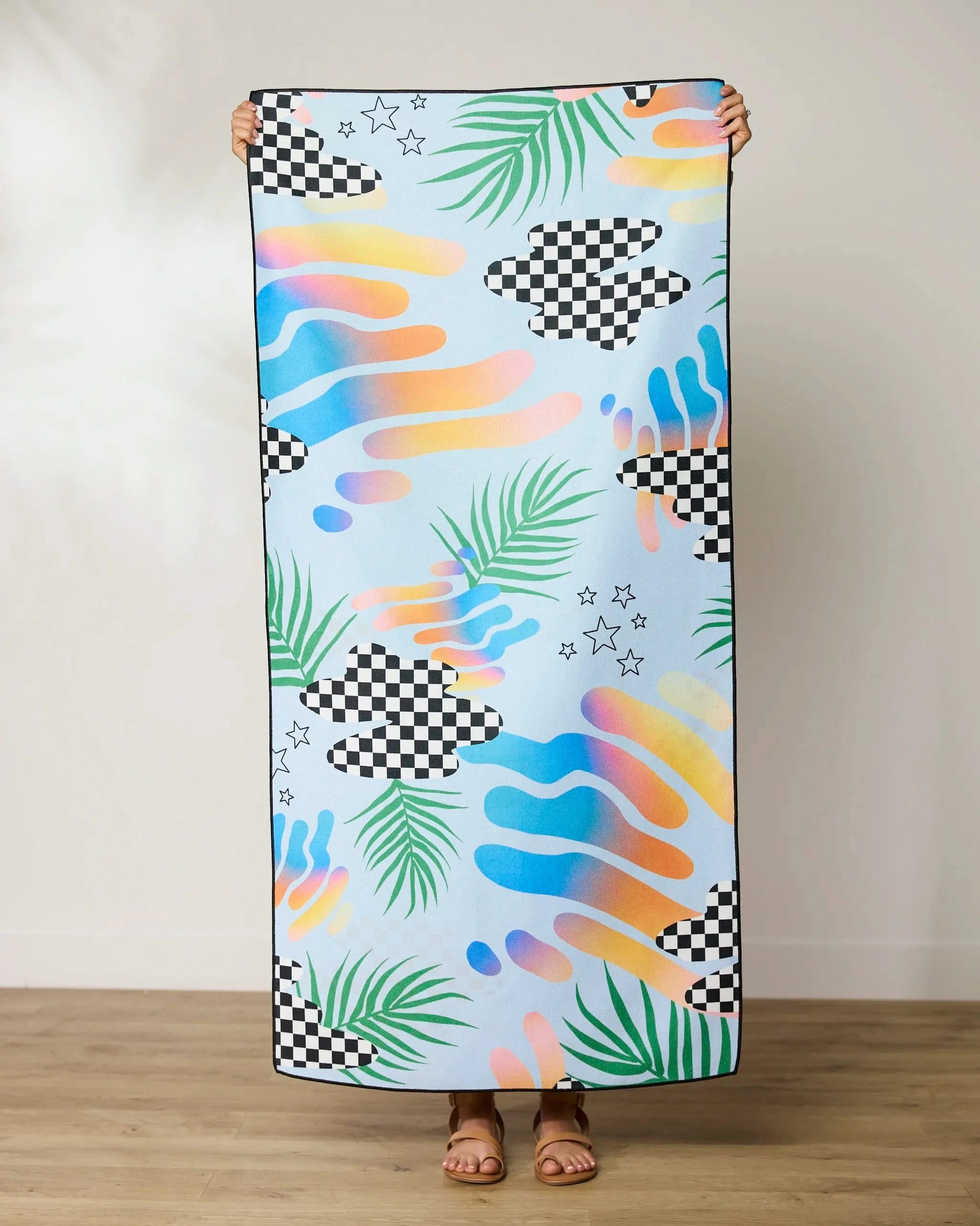 Someone holds up the GEOMETRY Playful Paradise Beach Towel, featuring colorful checkerboard patterns, pastel swirls, green palm leaves, and black stars—a vibrant, quick-drying towel made from recycled materials.