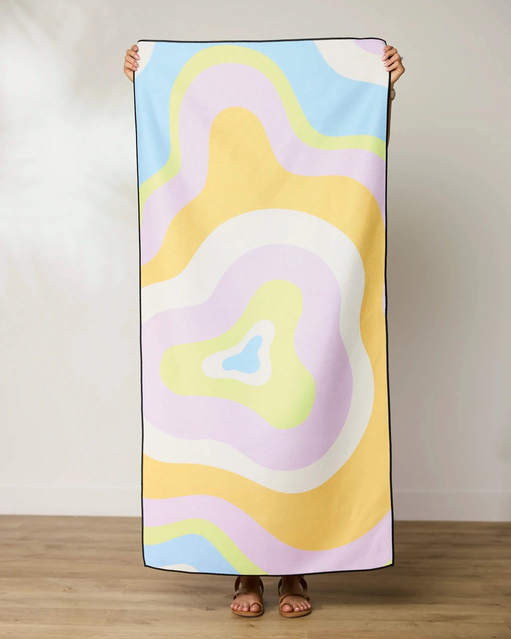 A person stands on a wooden floor holding up a GEOMETRY Swirls Beach Towel with abstract pastel shapes in yellow, purple, blue, green, and white. Their head and hands appear above this quick-drying towel.
