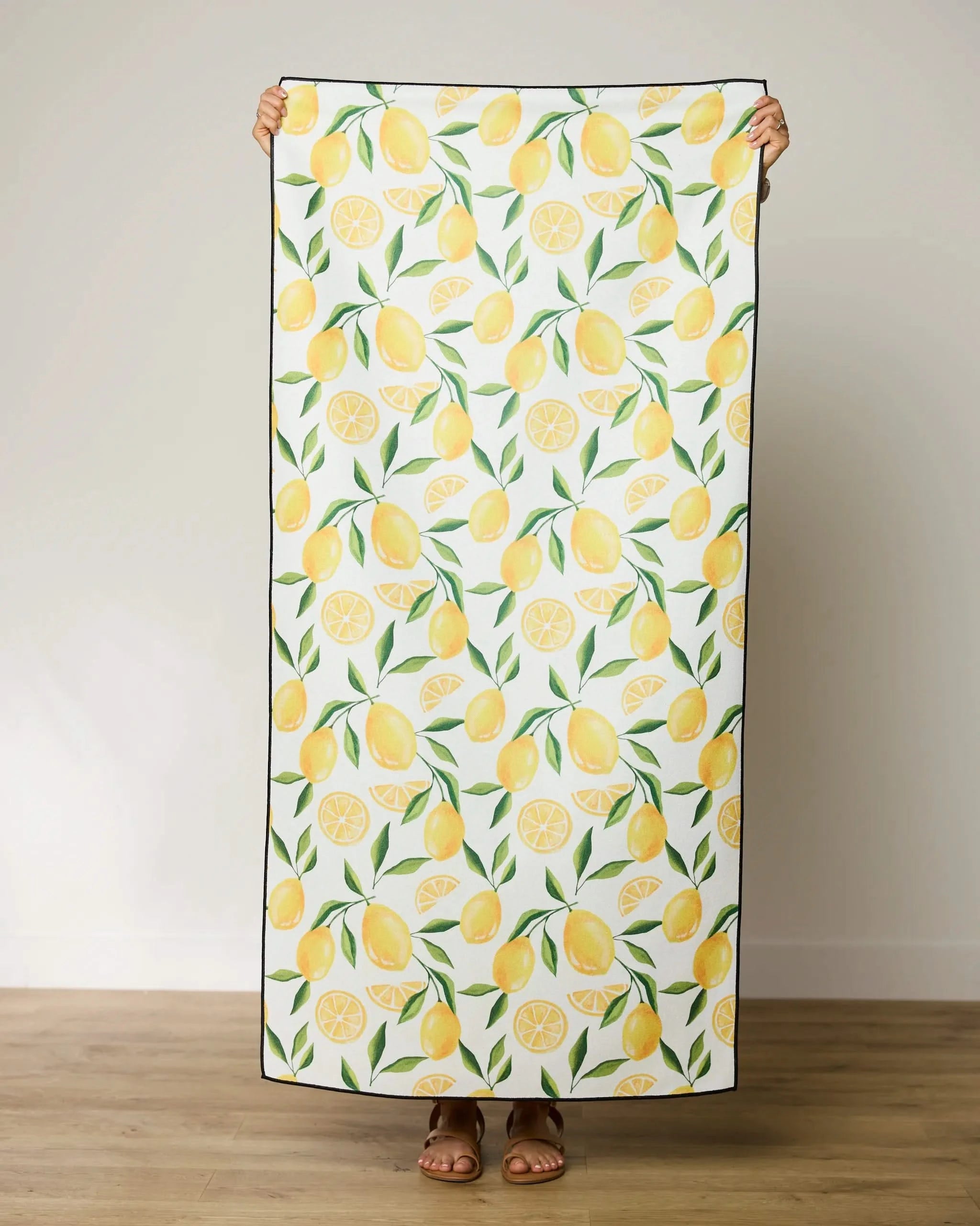 A person holds up the GEOMETRY Limoncello quick-drying micro-loop beach towel—white with whole lemons, slices, and green leaves. Only their hands, feet, and sandals are visible. Made from recycled materials for eco-friendly style.