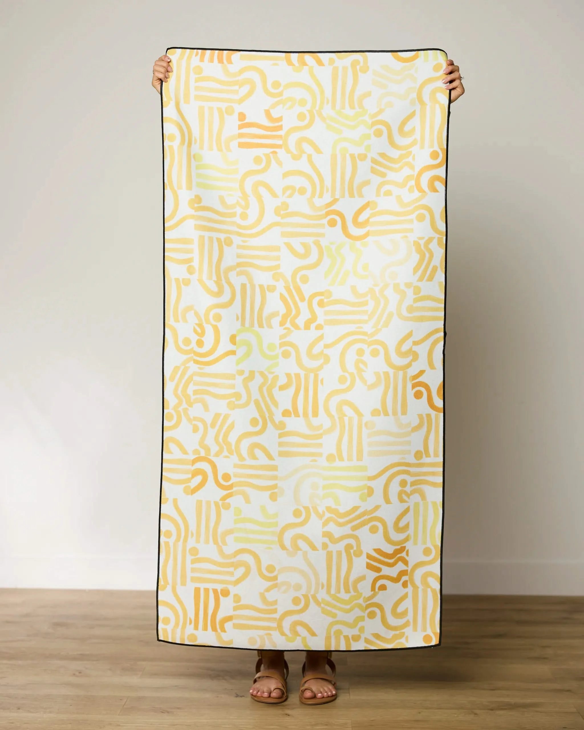 A person on a wooden floor holds up the GEOMETRY Venus Beach Towel with a yellow abstract line pattern on white; only their hands and feet are visible.