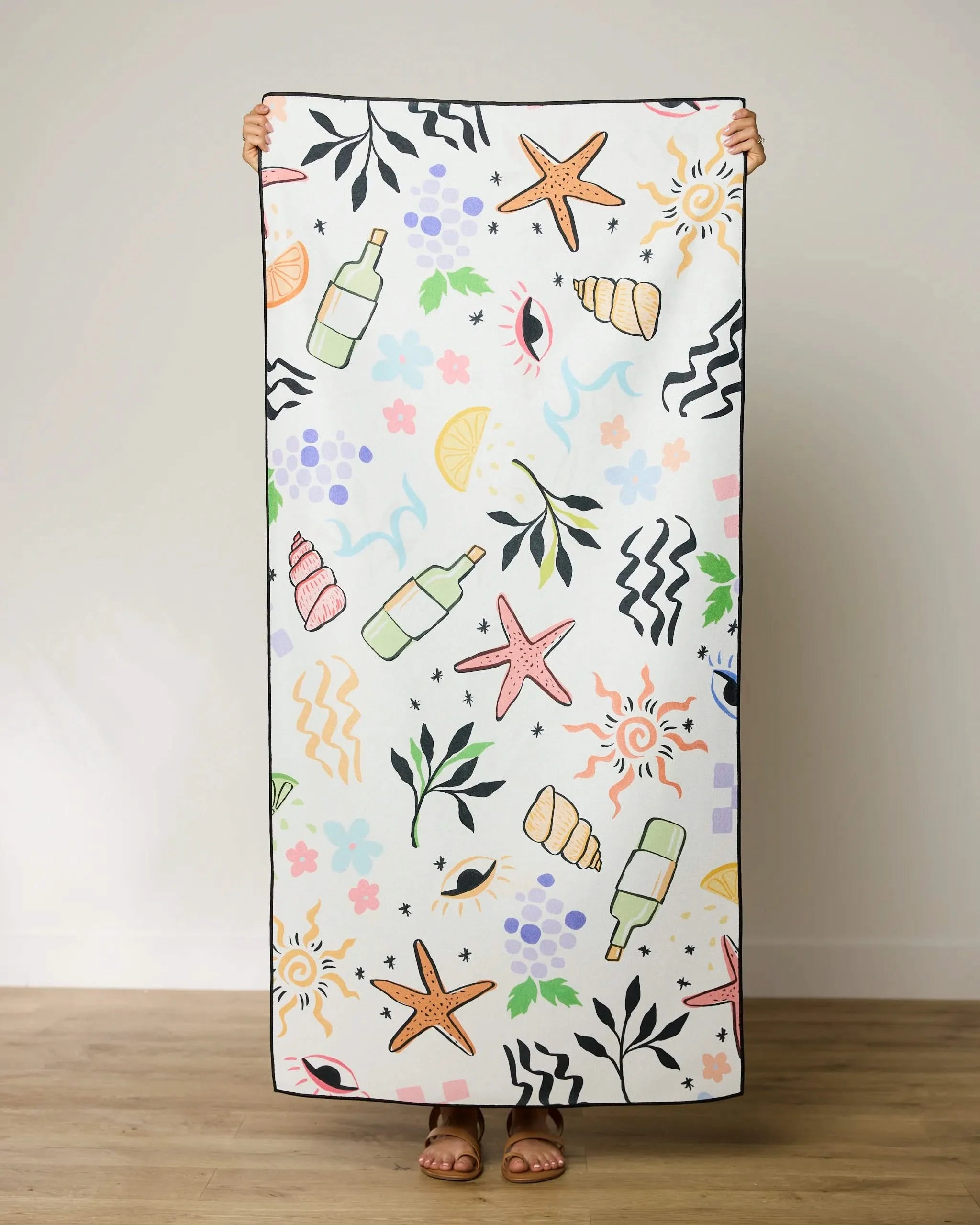 A person stands indoors holding up a GEOMETRY Wine Not beach towel featuring colorful illustrations of starfish, shells, bottles, leaves, fruits, flowers, waves, and abstract shapes. Only the persons feet are visible.