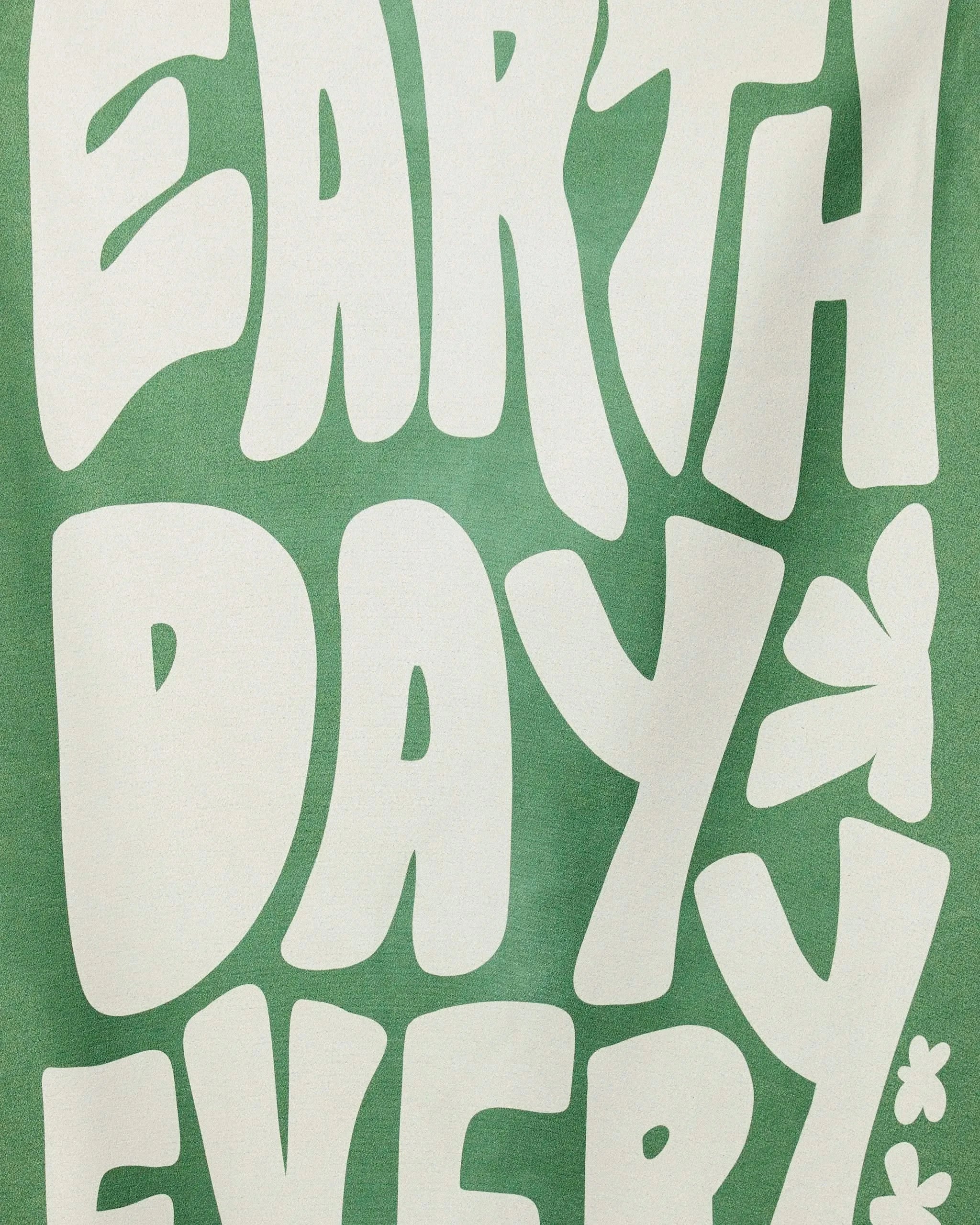 White bold letters on green spell “EARTH DAY EVERY” in playful, wavy fonts, with a small flower and butterfly—just like the fun patterns on the GEOMETRY Planet Pal Micro-loop Beach Towel made from recycled materials.
