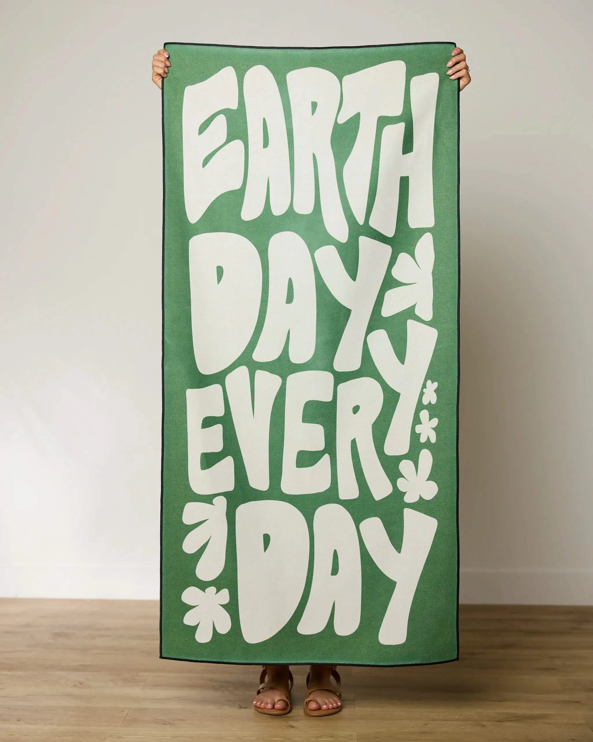 A person stands indoors holding a green EARTH DAY EVERY DAY sign with simple flower shapes—mirroring the eco-friendly style of GEOMETRY’s Planet Pal recycled materials beach towel.