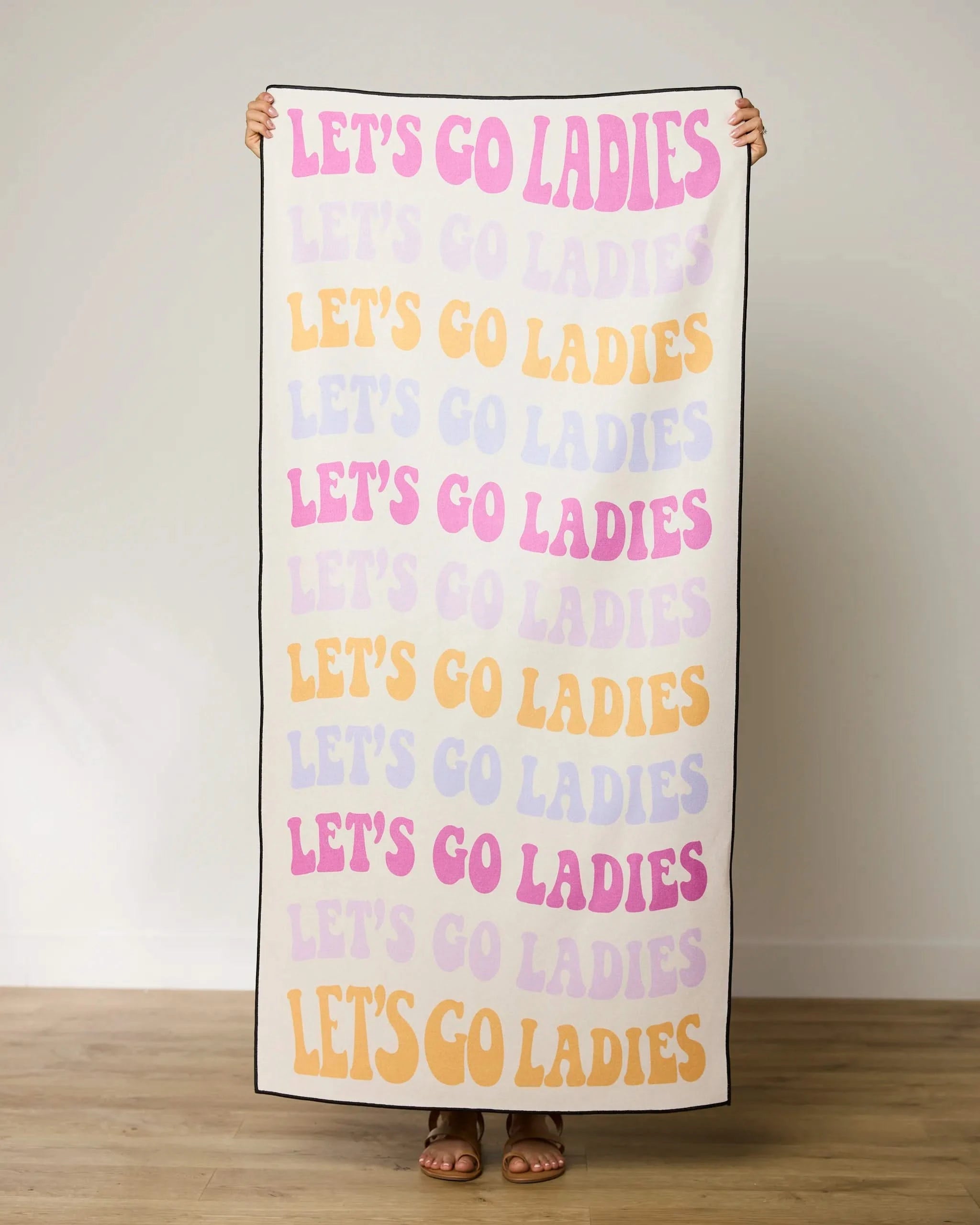 A person stands on a wooden floor holding GEOMETRYs Ladies Only Micro-loop Beach Towel, which features “LET’S GO LADIES” in pink, yellow, and lavender. The quick-drying towel is made from recycled materials; their feet peek beneath it.