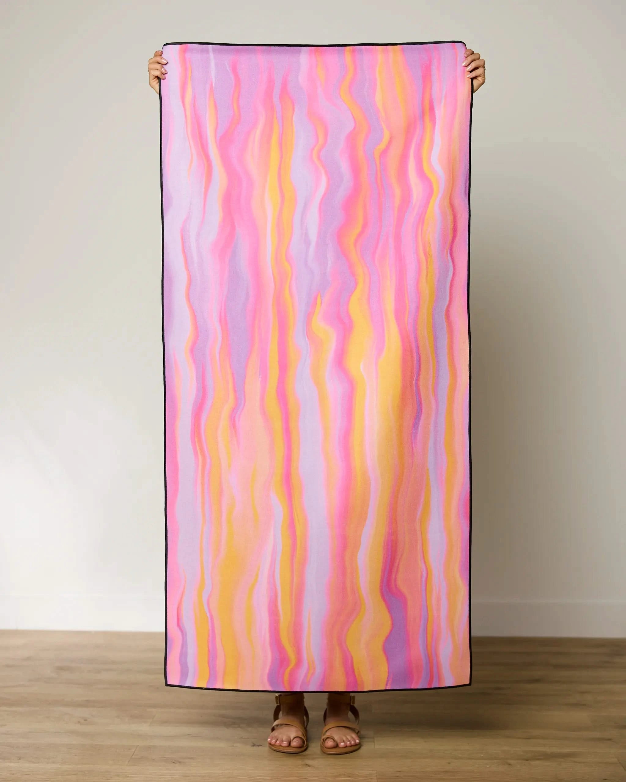 A person holds GEOMETRYs The Rush beach towel, featuring vertical pink, purple, yellow, and orange wavy stripes. Standing by a plain wall and wooden floor, they showcase this quick-drying towel made from recycled materials.