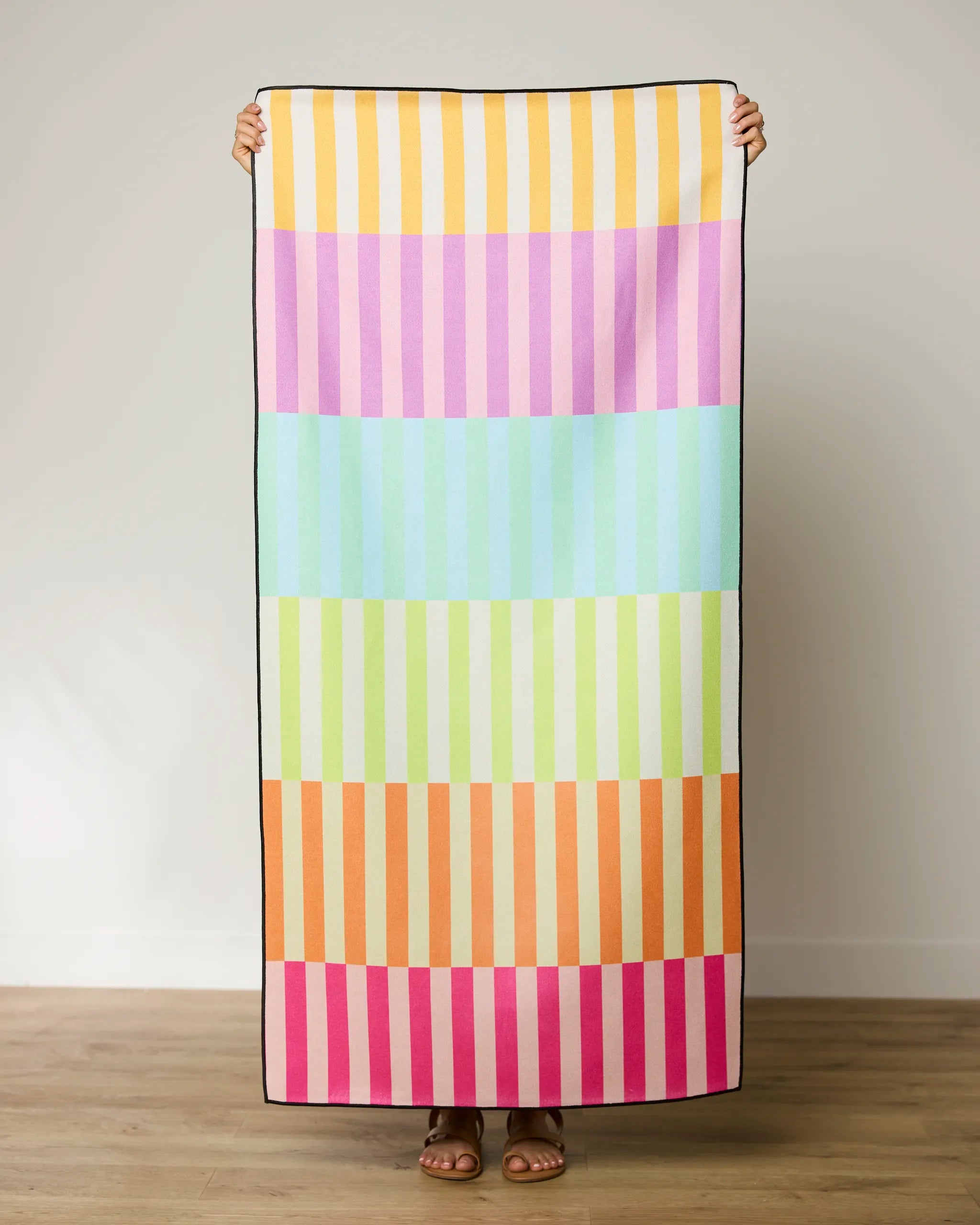 Someone holds up the GEOMETRY Neon Nights micro-loop beach towel, featuring horizontal stripes in pastel yellow, pink, blue, green, orange, and magenta. Made from recycled materials and quick-drying, its shown against a light wall and wooden floor.