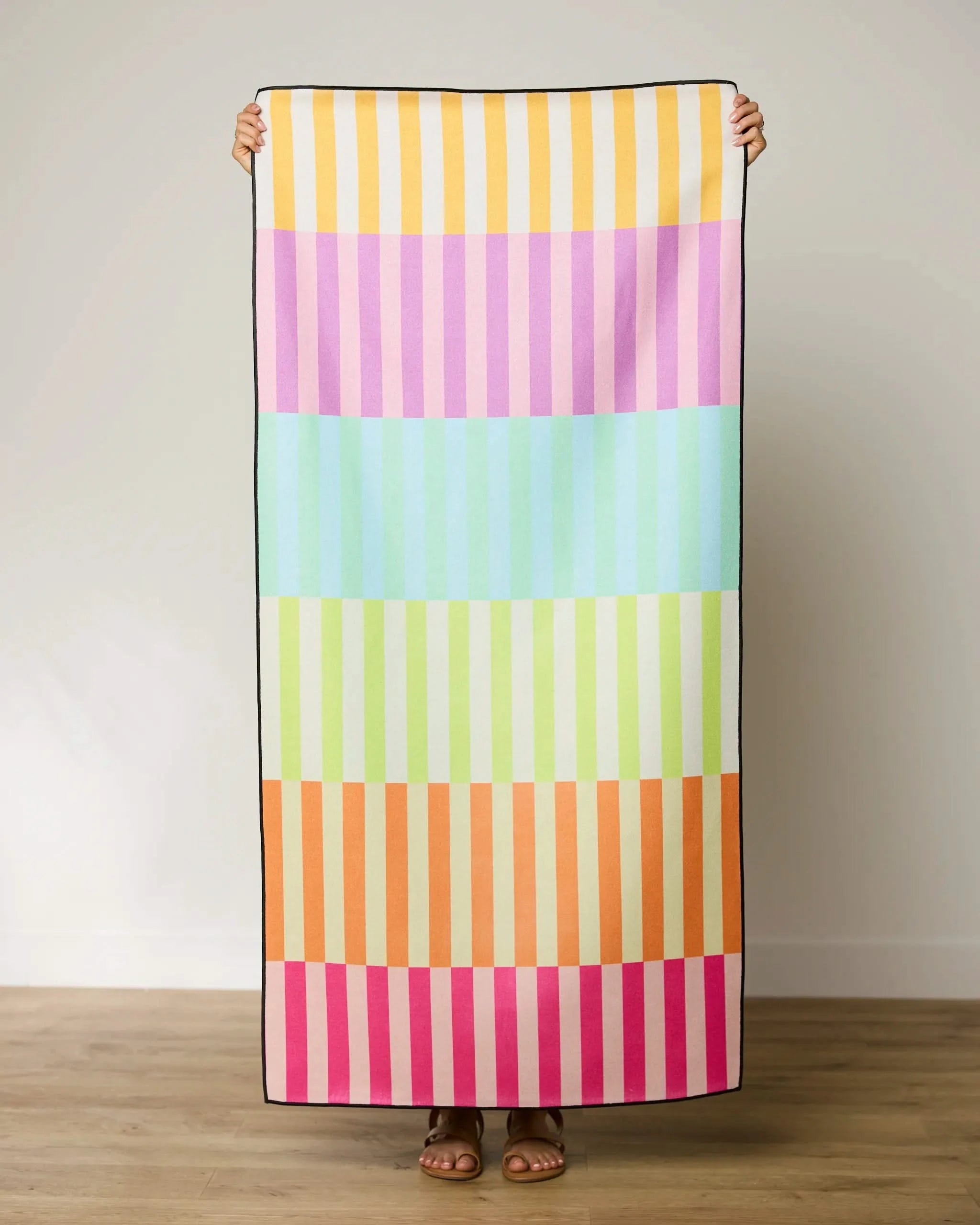 Someone holds up the GEOMETRY Neon Nights micro-loop beach towel, featuring horizontal stripes in pastel yellow, pink, blue, green, orange, and magenta. Made from recycled materials and quick-drying, its shown against a light wall and wooden floor.