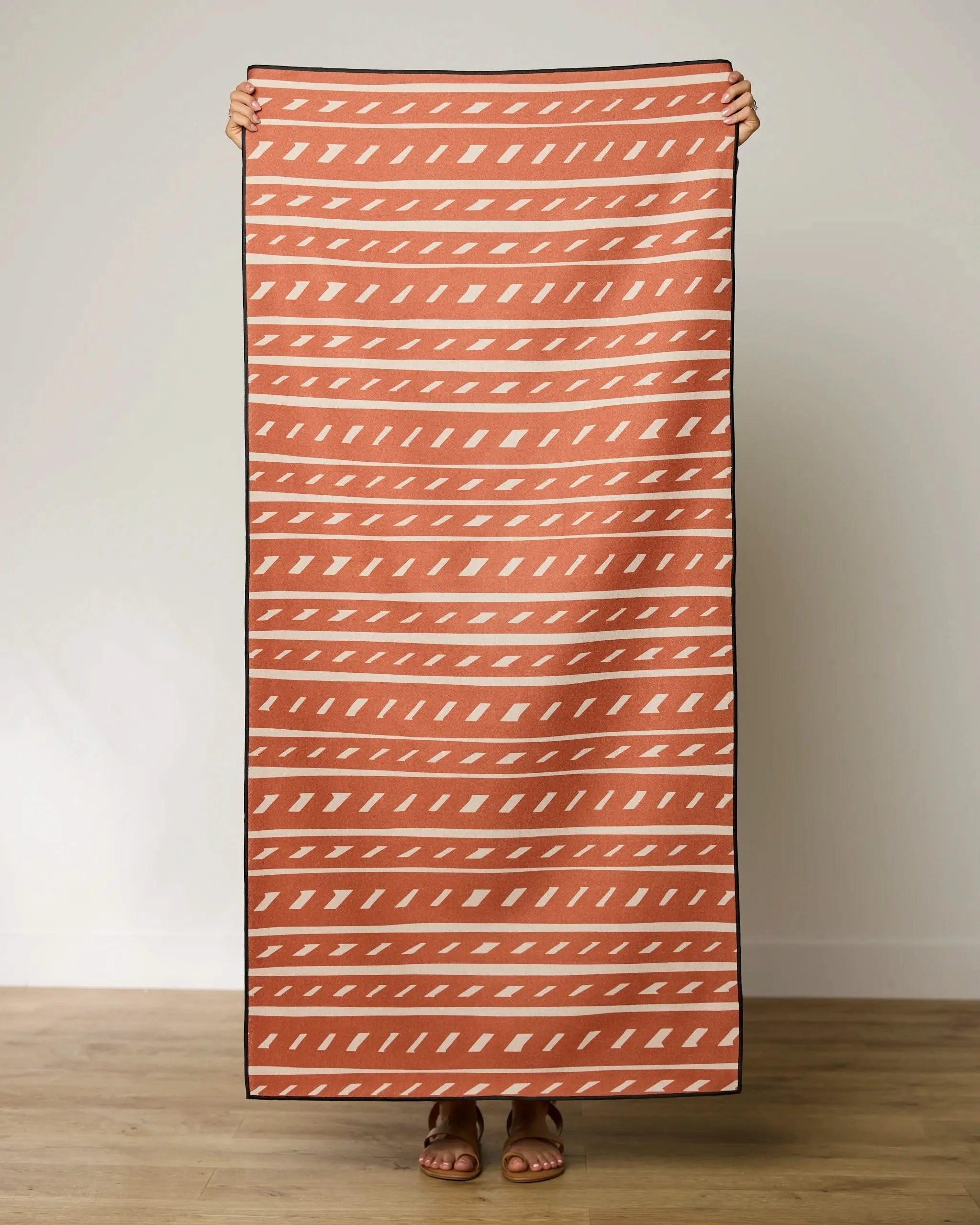 A person stands on a wooden floor, holding up a large GEOMETRY Auburn Autumn towel with white geometric diagonal lines and shapes; only their hands and feet are visible.
