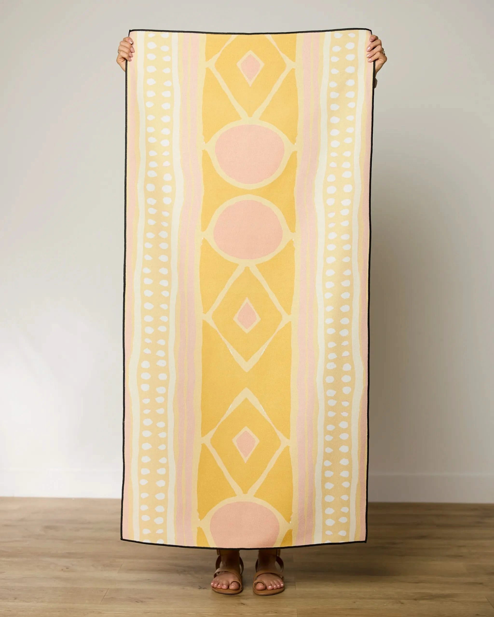 A person stands indoors holding up the GEOMETRY Beaded quick-drying beach towel in yellow, peach, and white geometric patterns, with only their hands and sandaled feet visible behind it.