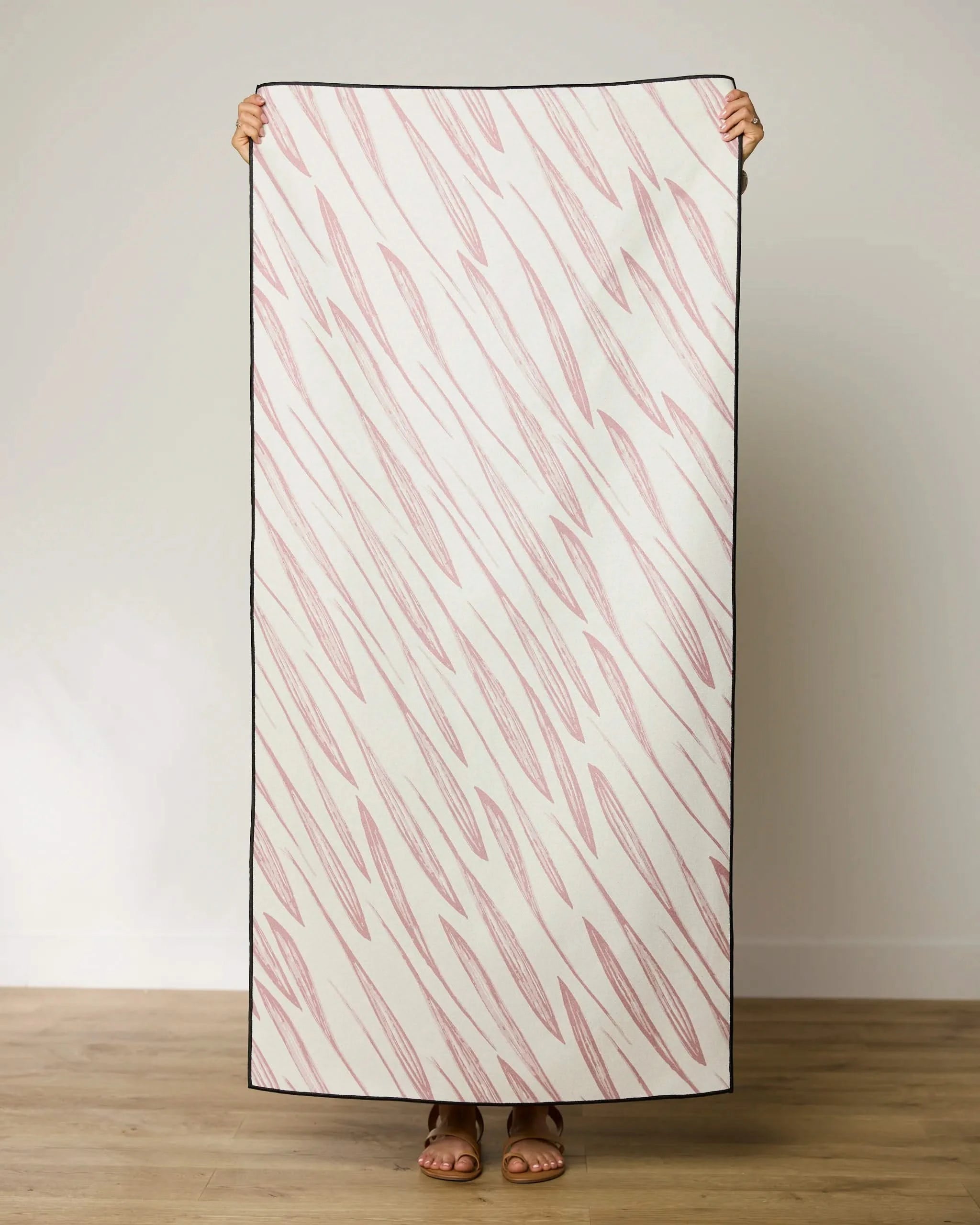 A person holds up the GEOMETRY Calm Winds Beach Towel, a rectangular quick-drying towel made from recycled materials, featuring a white background with diagonal pink zigzag lines. Only their hands and feet are visible behind the towel.