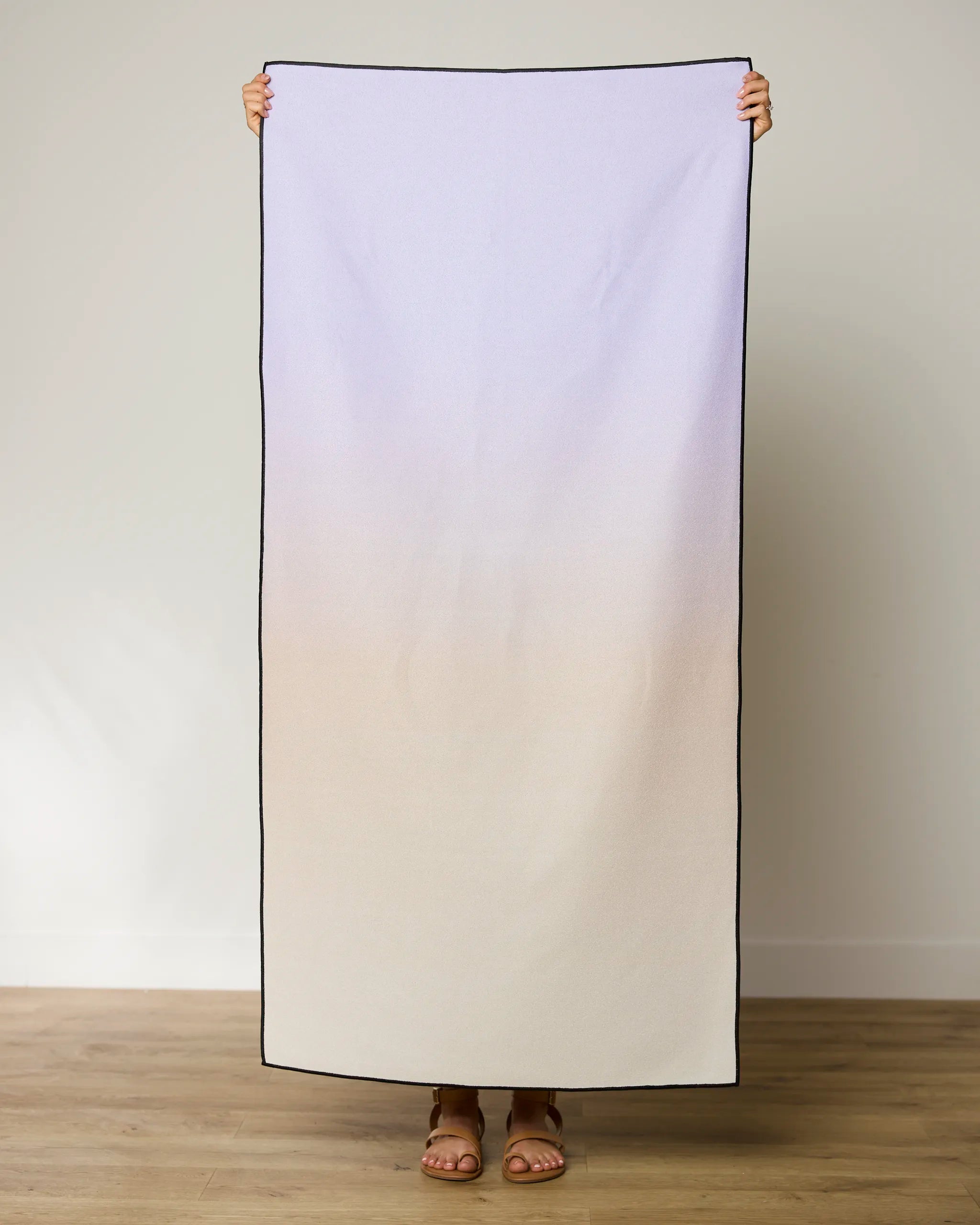 A person stands on a wooden floor holding up the GEOMETRY Groovy Gradient Micro-loop Beach Towel, showcasing its pale purple to beige gradient with black borders. Their feet in sandals peek out below this quick-drying towel made from recycled materials.