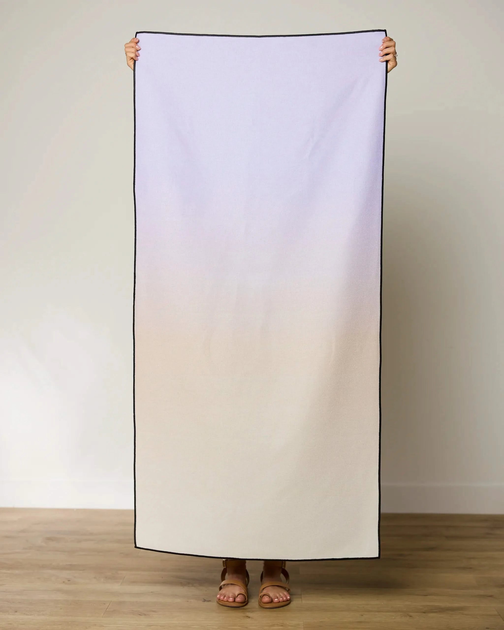 A person stands on a wooden floor holding up the GEOMETRY Groovy Gradient Micro-loop Beach Towel, showcasing its pale purple to beige gradient with black borders. Their feet in sandals peek out below this quick-drying towel made from recycled materials.