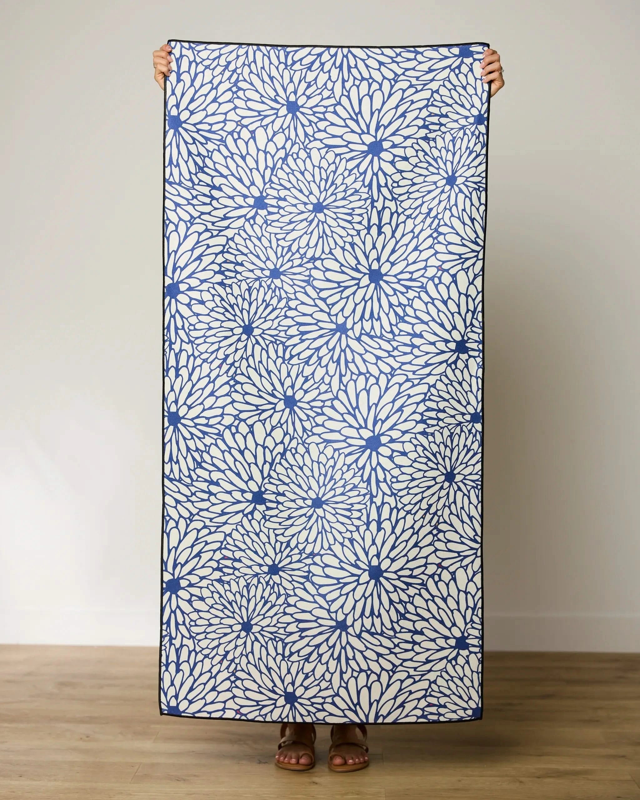 A person holds up a rectangular, blue and white floral GEOMETRY Jar Jar Micro-loop Beach Towel indoors on a wooden floor against a plain light wall. Only their hands, feet, and sandals are visible.