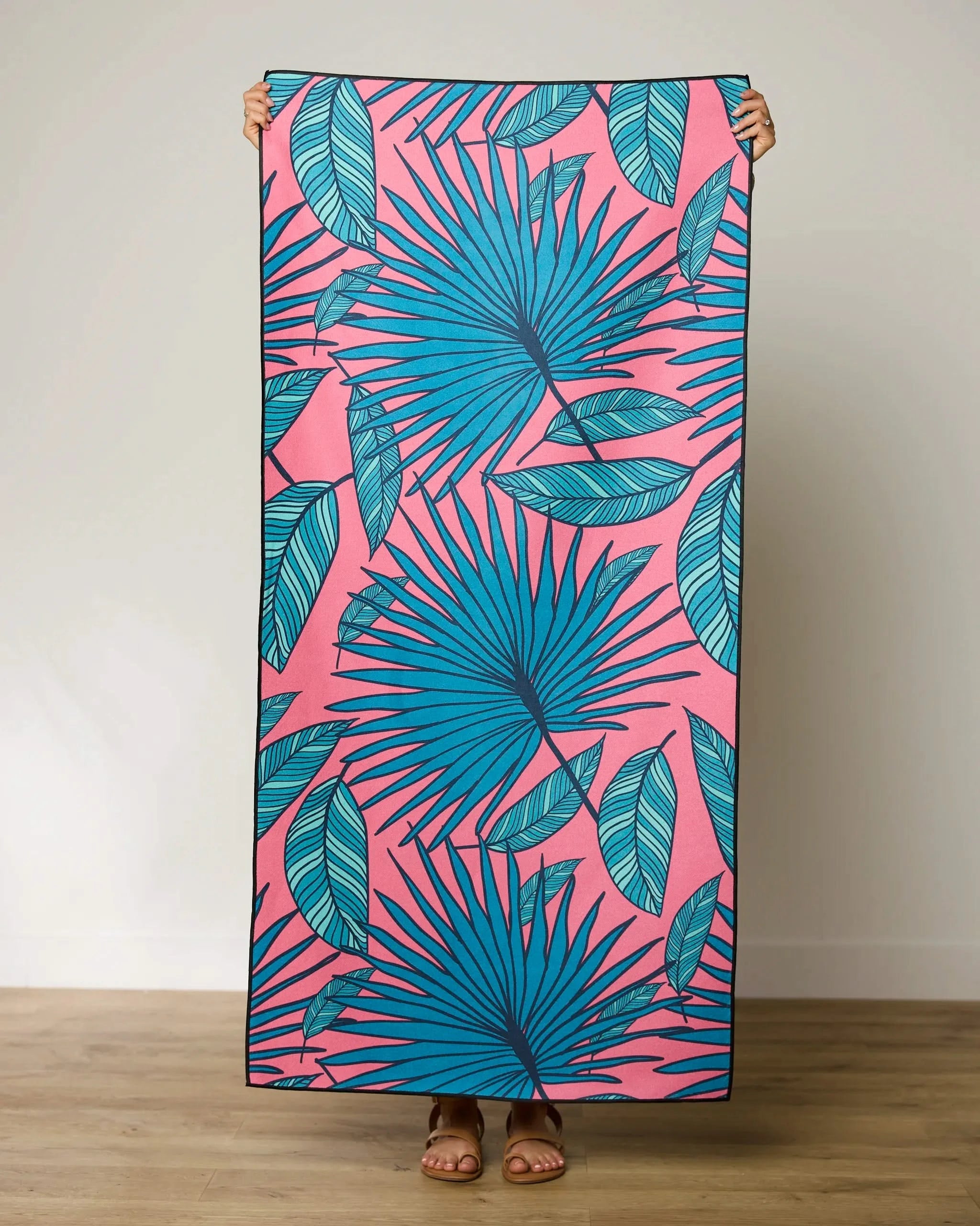 A person holds the GEOMETRY Jazzy Jungle micro-loop beach towel, featuring a pink background with bold blue tropical leaves. Only their hands and feet are visible indoors on a wooden floor. This quick-drying towel adds stylish flair anywhere.
