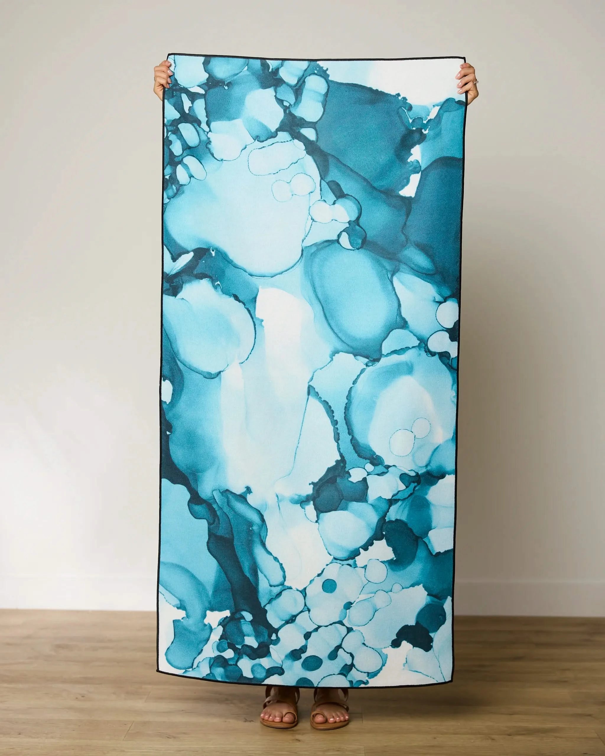 A person stands indoors holding the GEOMETRY Leah beach towel, featuring a large, quick-drying design with a blue and teal abstract pattern, covering their body except for their sandal-clad feet.
