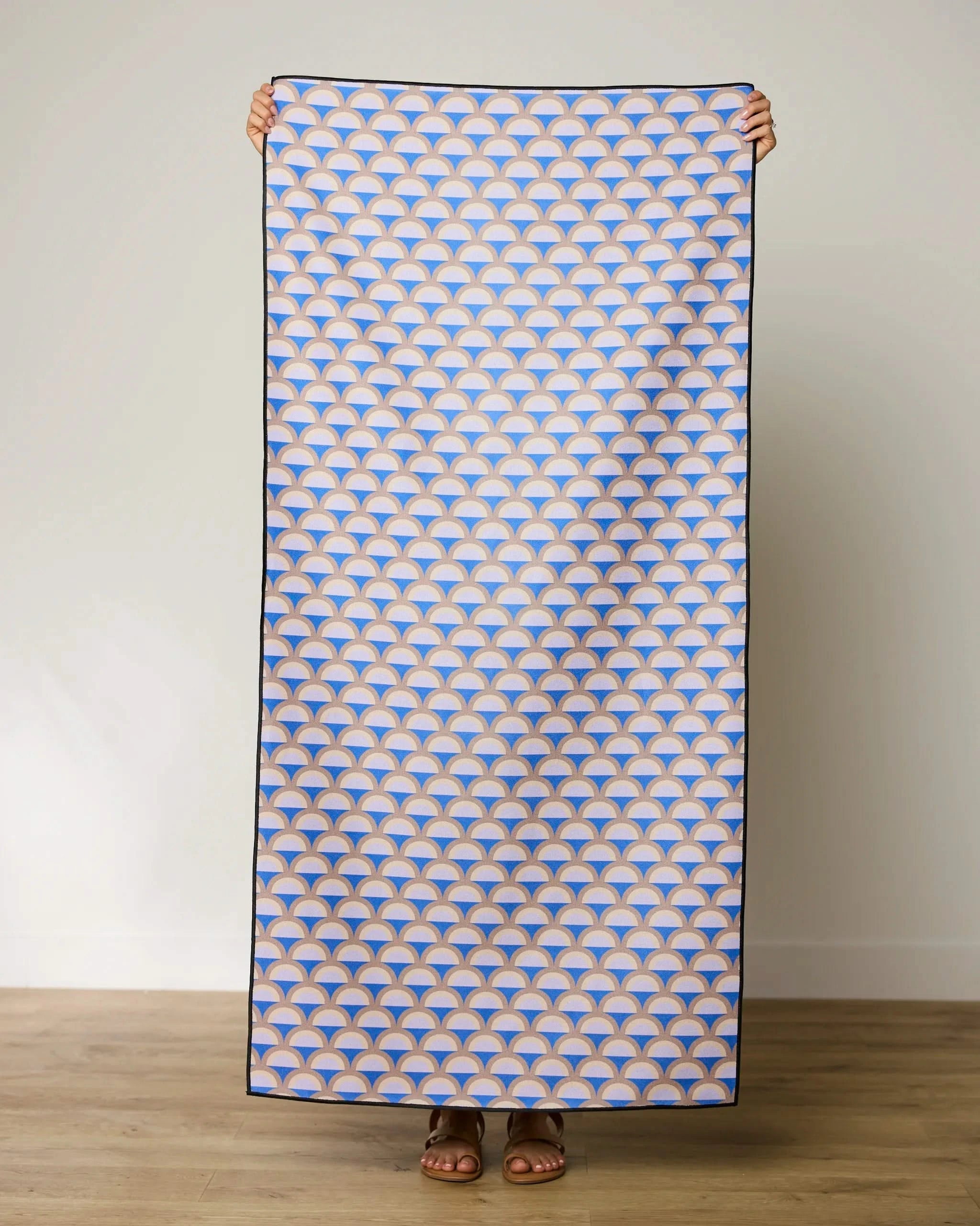 A person stands holding up the GEOMETRY Mauve Mounds quick-drying beach towel, which features a repeating blue, orange, and white geometric fan pattern, against a plain light wall and wooden floor.