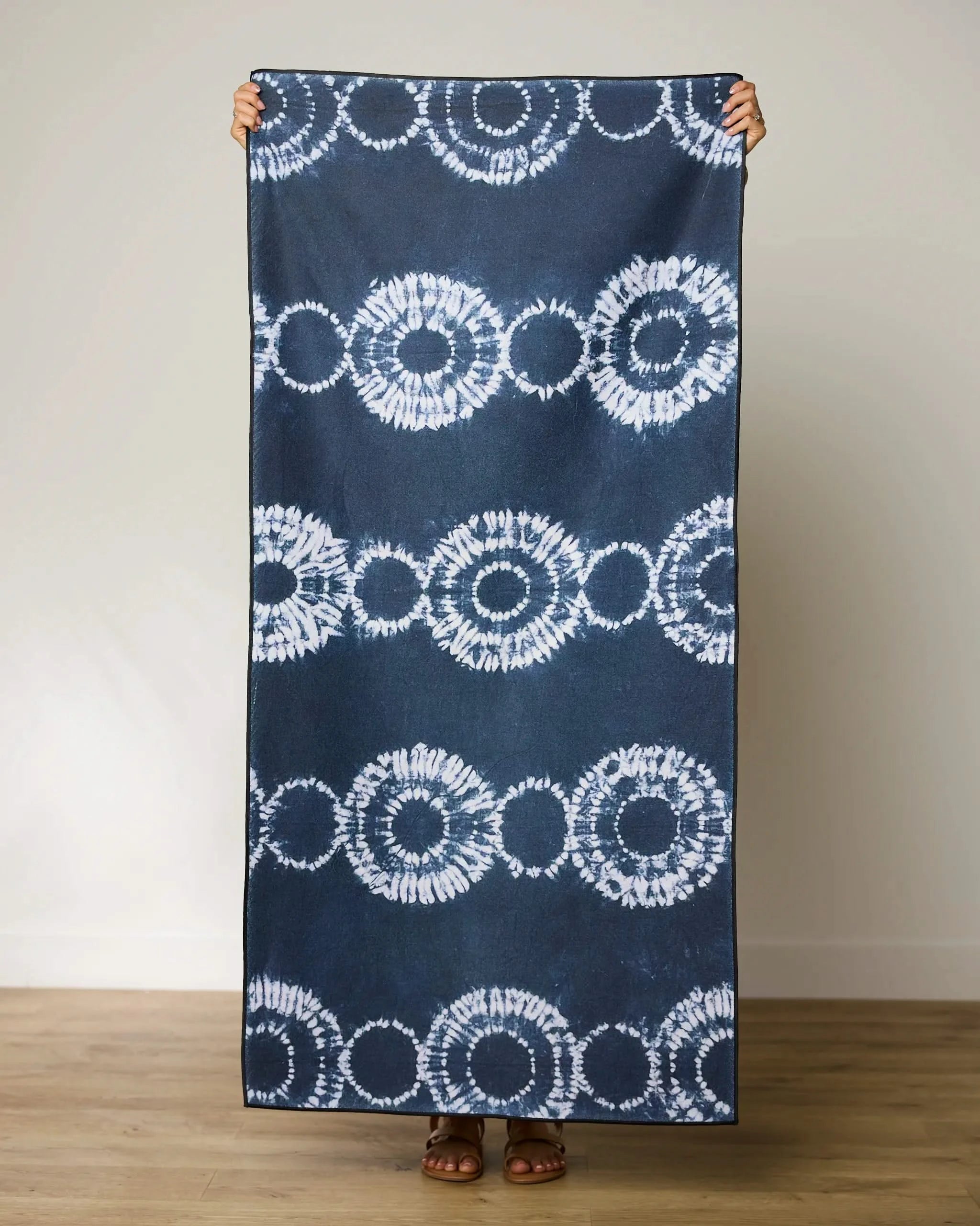 A person stands on a wooden floor holding up the GEOMETRY Shibo Stars large blue and white micro-loop beach towel made from recycled materials, covering their body except hands and feet, against a plain light wall.