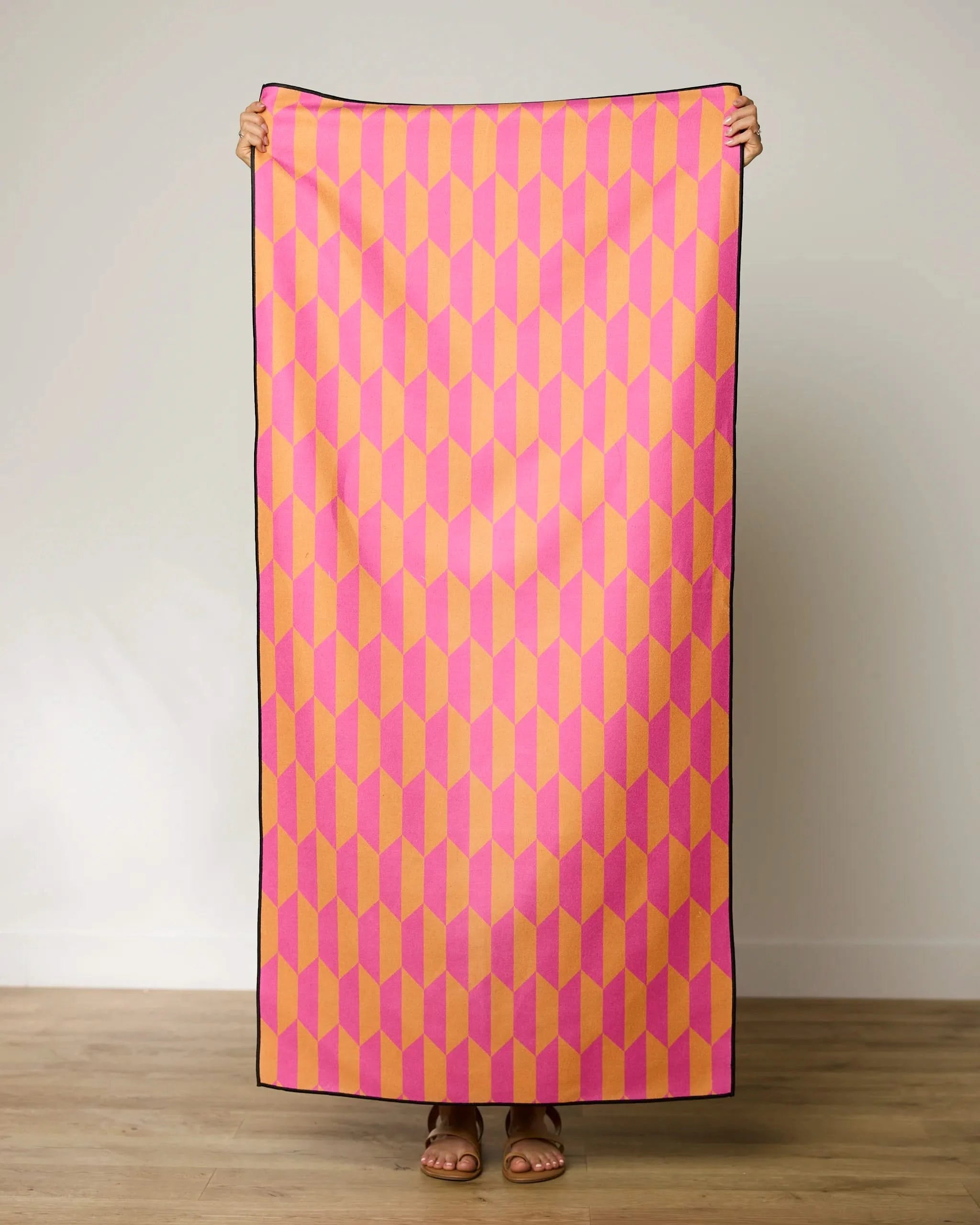 A person stands indoors holding up the GEOMETRY Bright Trapezoid beach towel, featuring a pink, yellow, and orange geometric pattern; only their hands, feet, and sandals are visible.