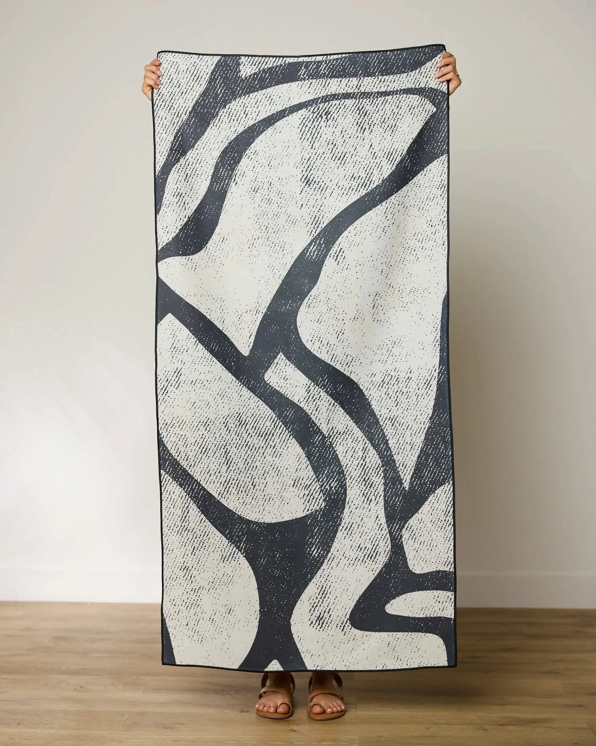 A person holds up the GEOMETRY Block Micro-loop Beach Towel, featuring an abstract black and white rectangular design, hiding their face and body except for hands and brown sandals, set against a plain light background.
