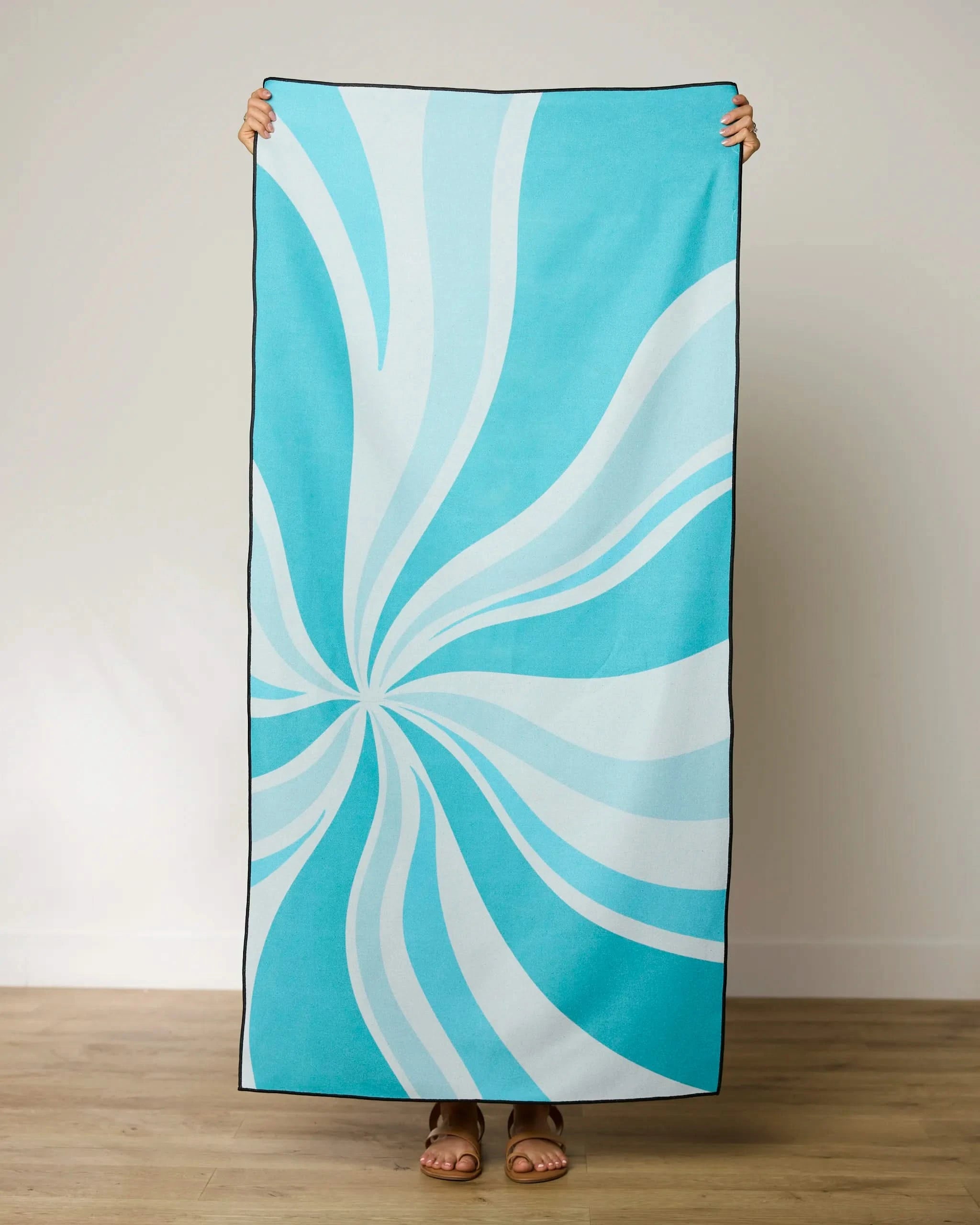 A person stands indoors on a wooden floor, holding up the GEOMETRY Candy Blue beach towel with a swirling white pattern. Only their hands and feet are visible behind this stylish, quick-drying towel.