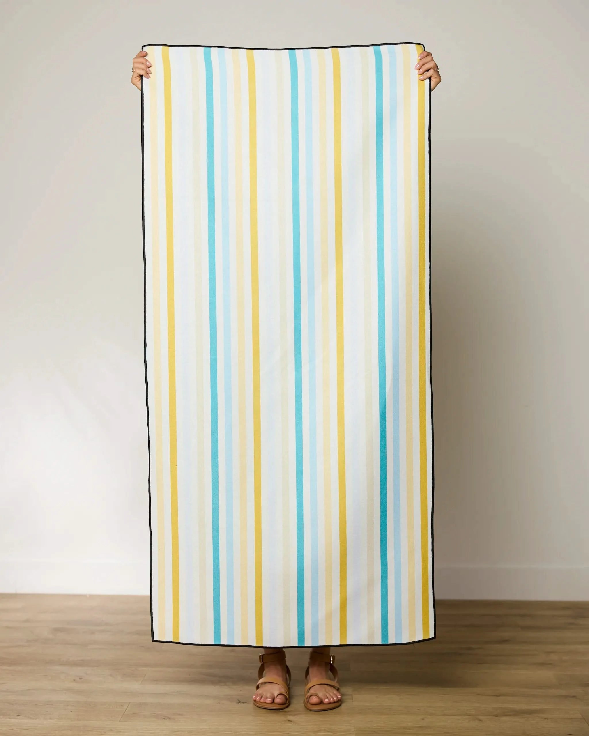 A person stands indoors holding up a GEOMETRY Seaside Sunshine beach towel with vertical blue, yellow, and white stripes; only their hands and sandal-clad feet are visible.