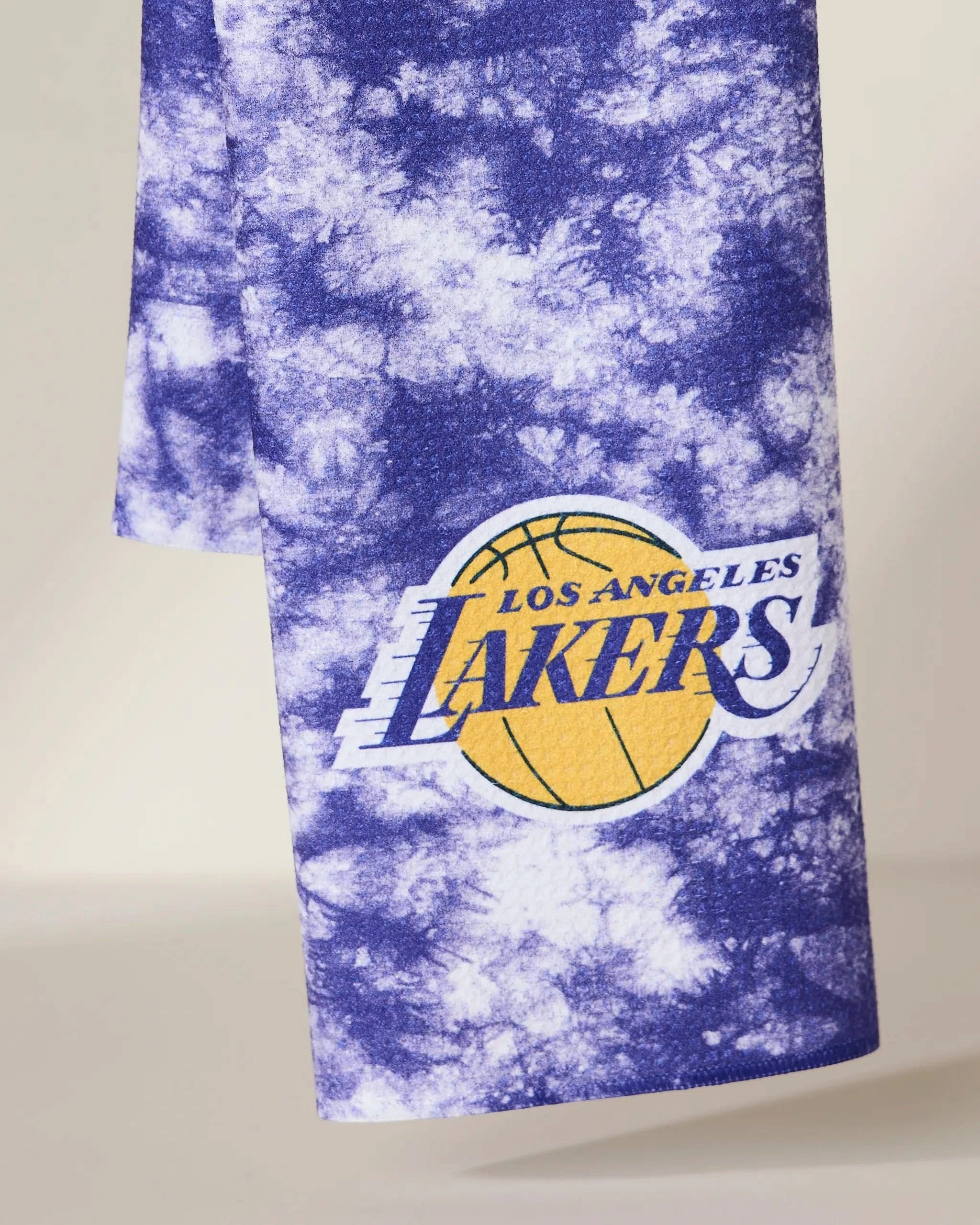 The Geometry Lakers Flash towel, featuring blue and white tie-dye with the yellow and purple Los Angeles Lakers logo and basketball graphic, is shown hanging against a neutral background.