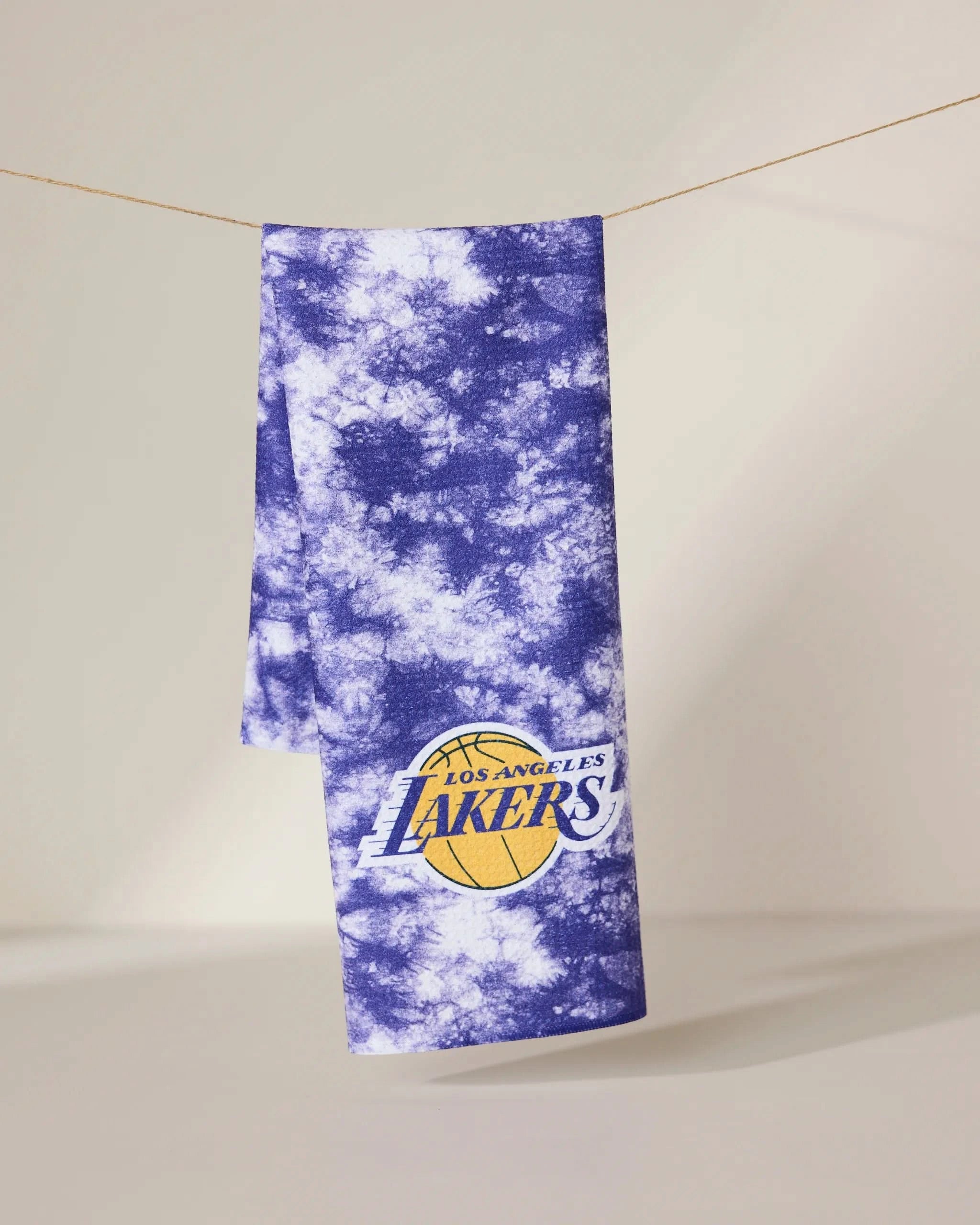 The Lakers Flash towel by Geometry, featuring a purple and white tie-dye design with the Los Angeles Lakers logo, hangs on a line against a plain beige background.