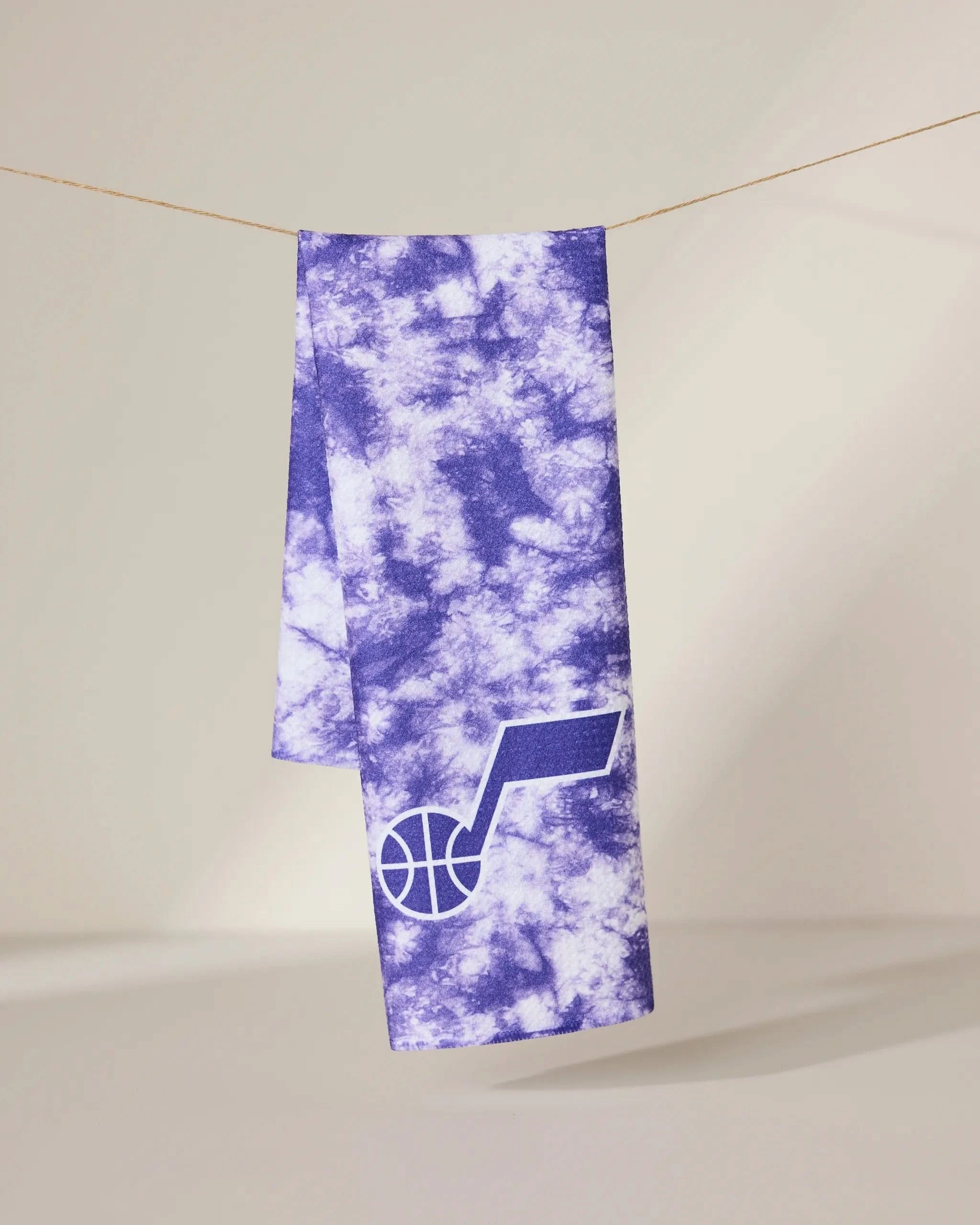 The Geometry Jazz Flash towel, featuring a purple and white tie-dye design with a basketball and music note logo on one end, hangs on a string against a neutral background.